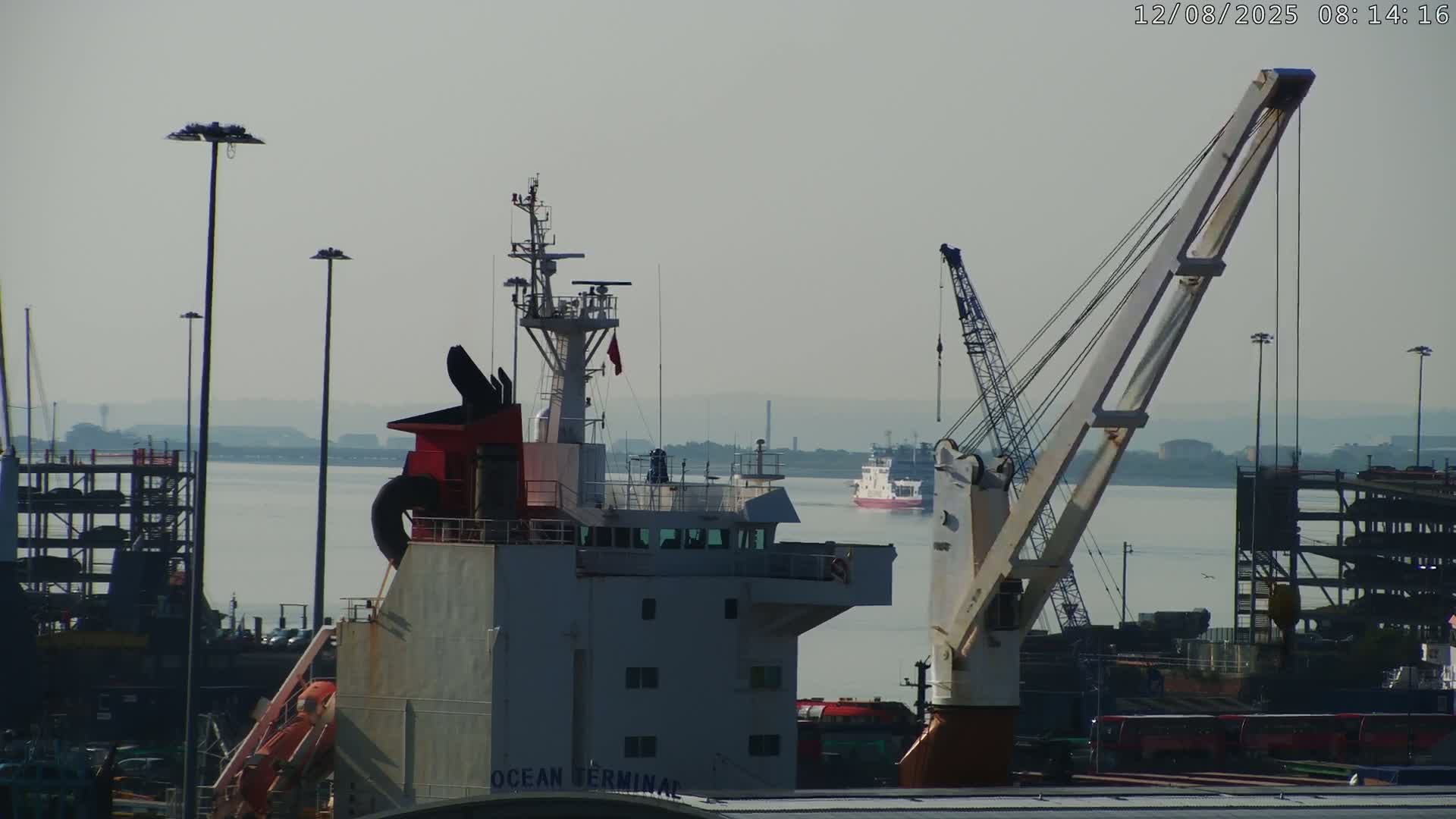 A hazy day at a port shows a large ship's bridge and a crane in the foreground, with another ship and industrial structures visible in the background across calm water.