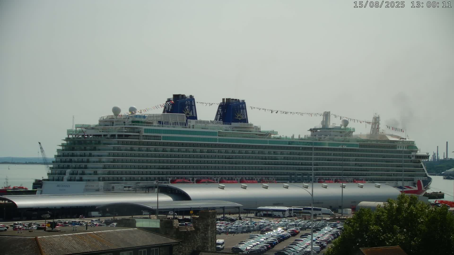 A hazy day at a port shows a large ship's bridge and a crane in the foreground, with another ship and industrial structures visible in the background across calm water.