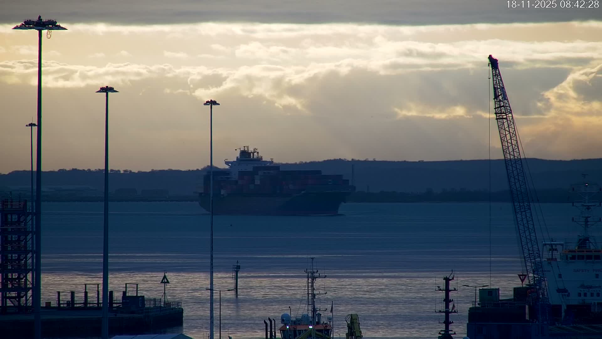 A large container ship sails on a body of water beneath a dramatic partly cloudy sky with sunlight breaking through, flanked by tall port light poles and a prominent construction crane in the foreground.