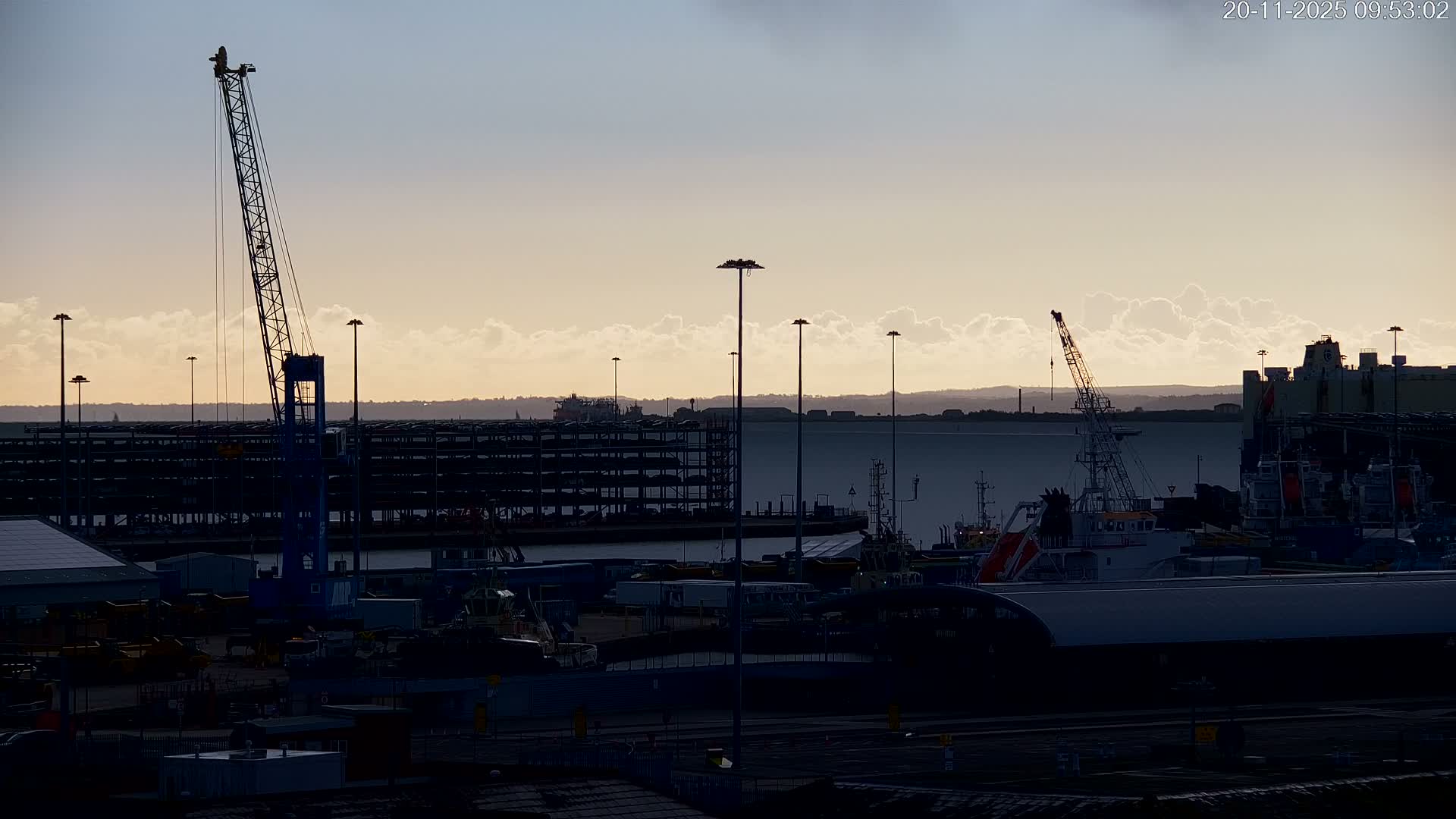 A bustling harbor is seen under a partly cloudy late morning sky, with multiple cranes, large industrial buildings, and various ships silhouetted against the bright, warm-toned horizon stretching over a calm body of water.