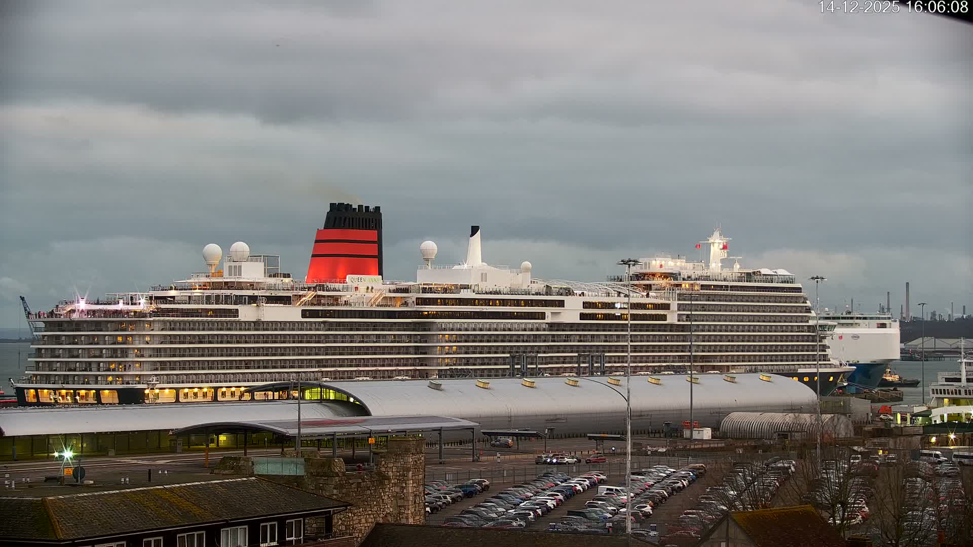 A large cruise ship, featuring a prominent Union Jack design on its bow, is docked at a port with a full parking lot in the foreground, under a cloudy morning sky with soft orange and pink hues suggesting sunrise.