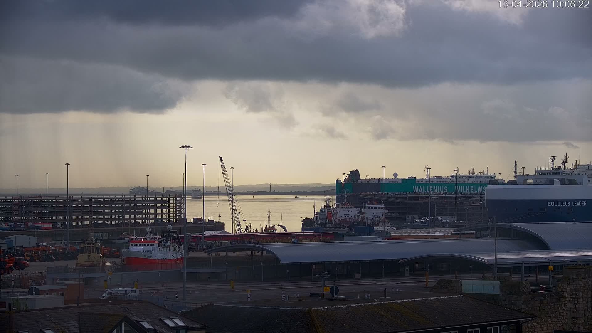 A large cruise ship, featuring a prominent Union Jack design on its bow, is docked at a port with a full parking lot in the foreground, under a cloudy morning sky with soft orange and pink hues suggesting sunrise.