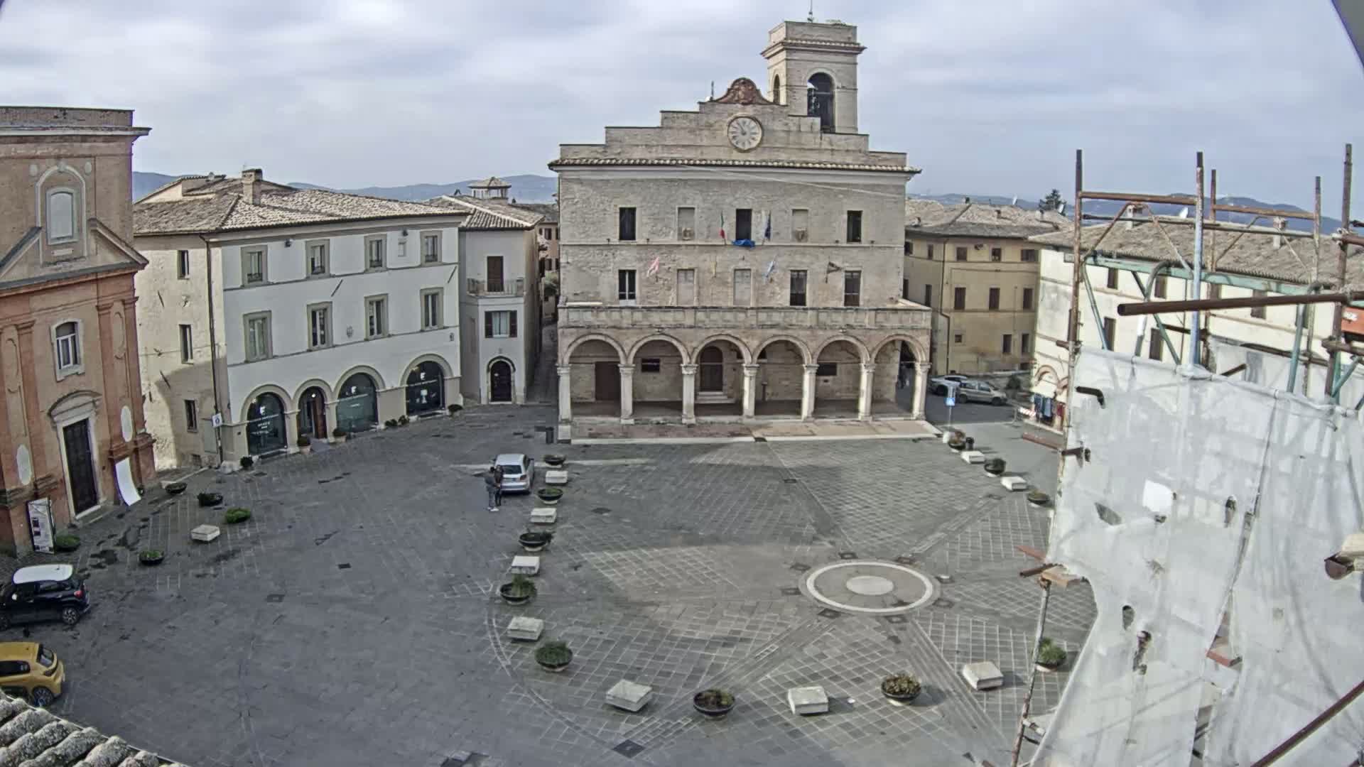 A large, mostly empty town square with a clock tower and several buildings, under a cloudy sky.