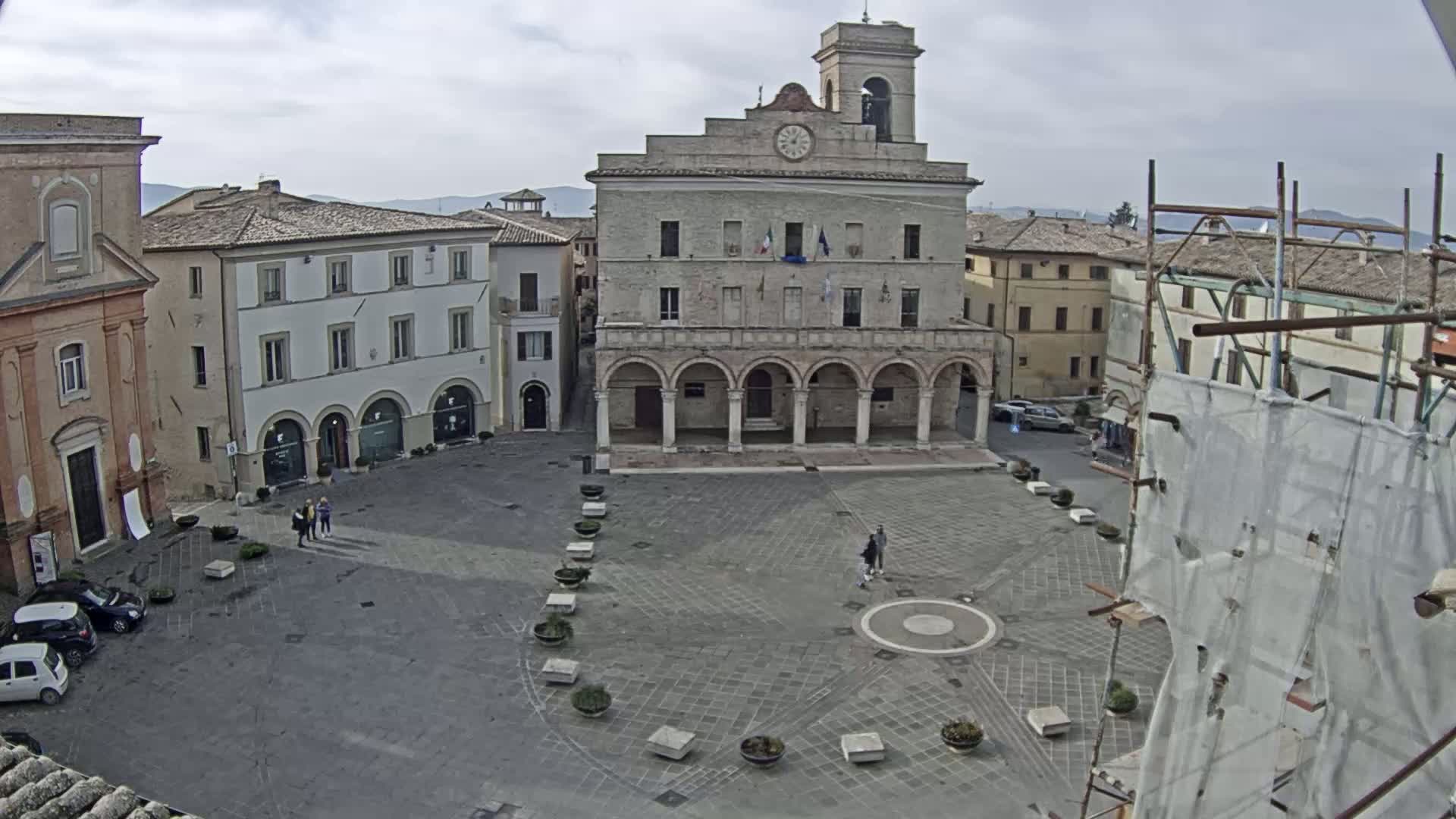 A large town square, under an overcast sky, features a prominent stone building with arched walkways, surrounded by other buildings and a few people.