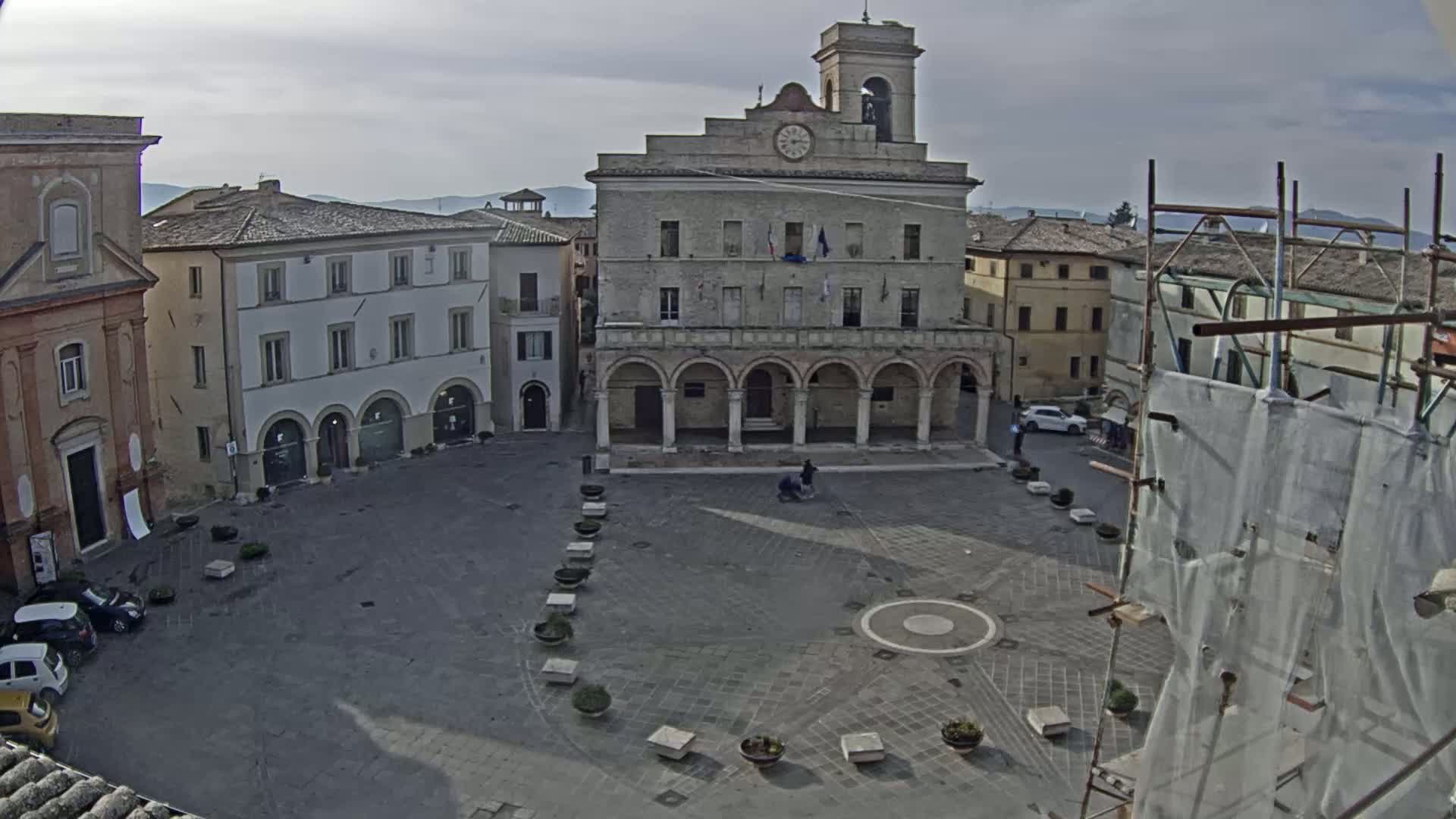 A town square with a large building featuring arched entrances and a clock tower, surrounded by other buildings under an overcast sky.