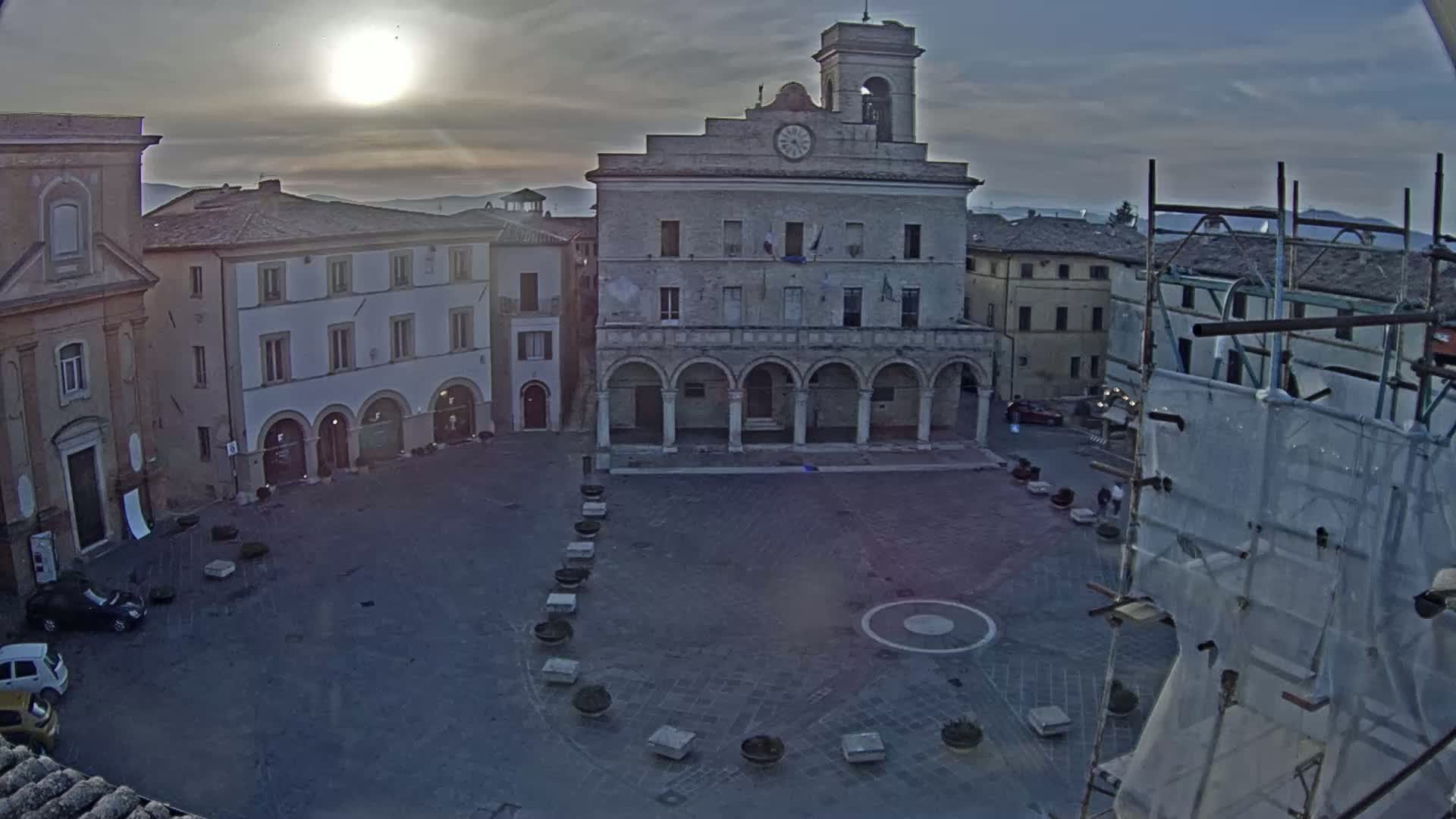 A mostly empty town square, under a partly cloudy sunset, is centered around a large, arched stone building with a clock tower.