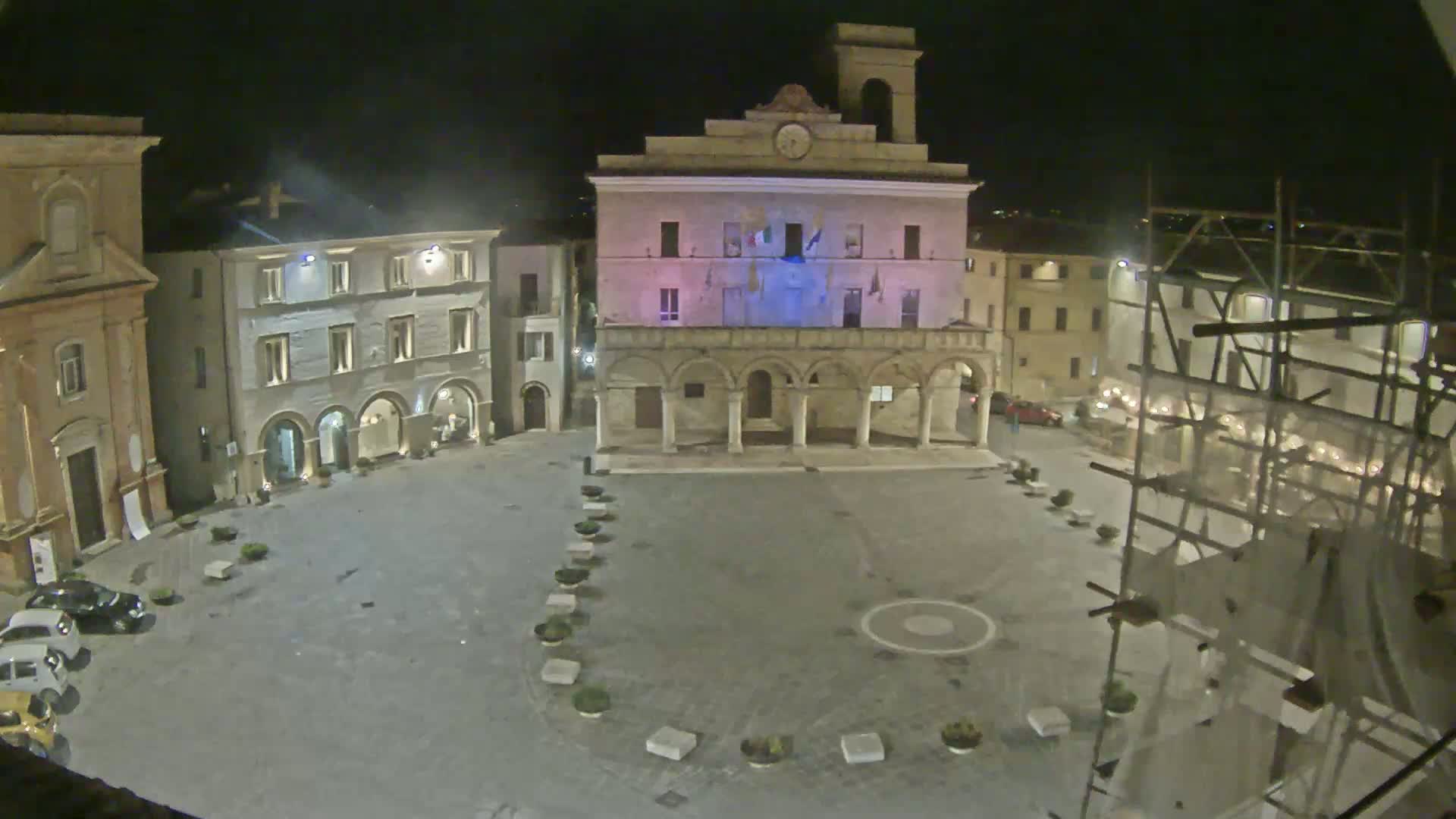 A nighttime view of an empty town square, with several buildings surrounding it, one of which is brightly lit.
