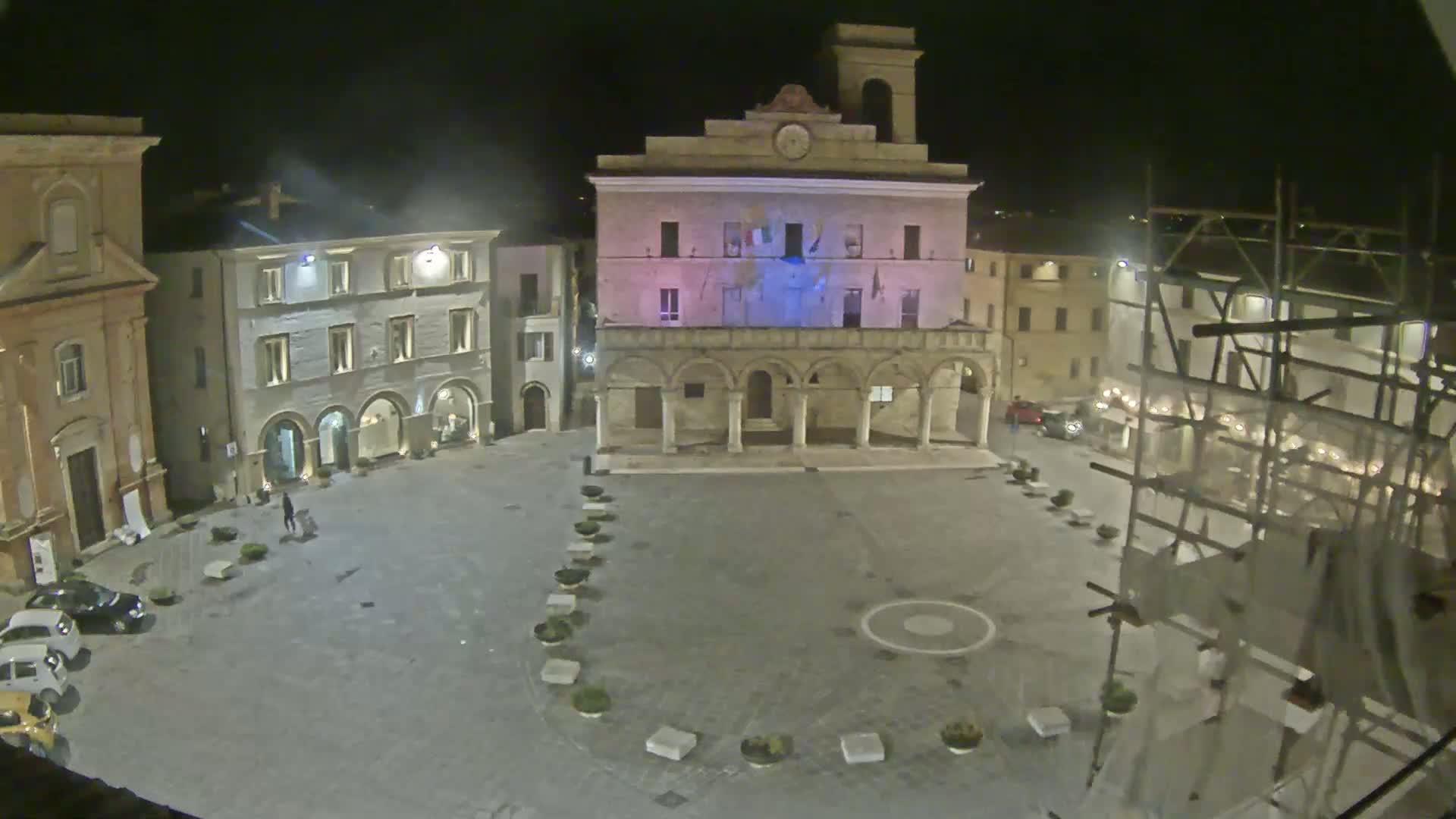 At night, an empty town square is illuminated, centered on a large building with arched entrances, surrounded by other buildings, some with scaffolding.