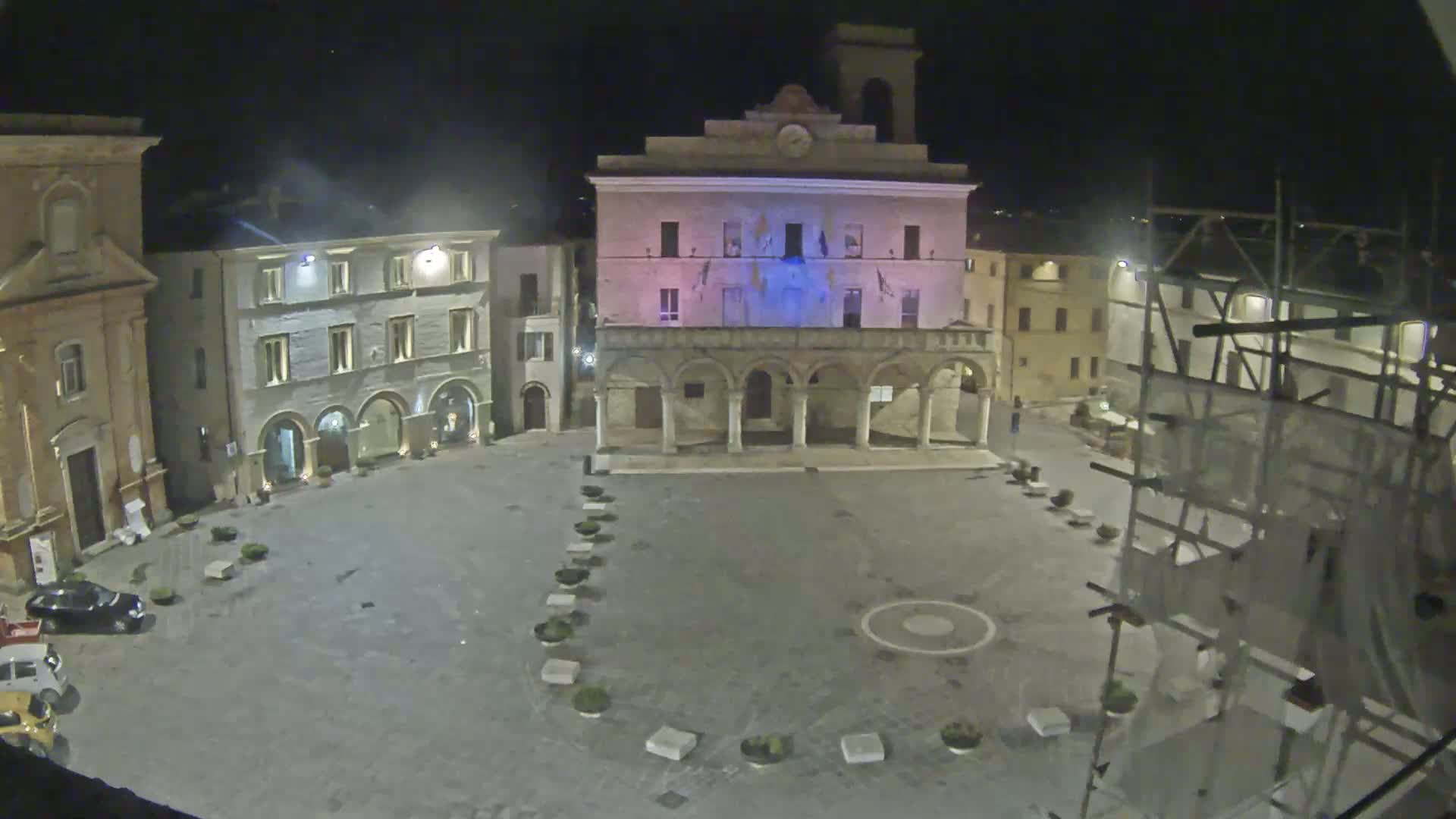 An empty, stone plaza at night is surrounded by historic buildings, one of which is brightly lit, with scaffolding visible near one building.