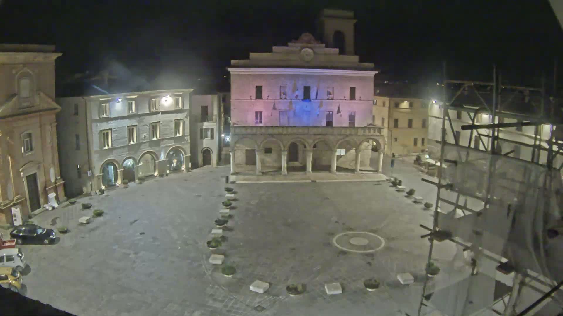 A nighttime view of an empty town square surrounded by historic buildings, one of which is illuminated in pink and blue.