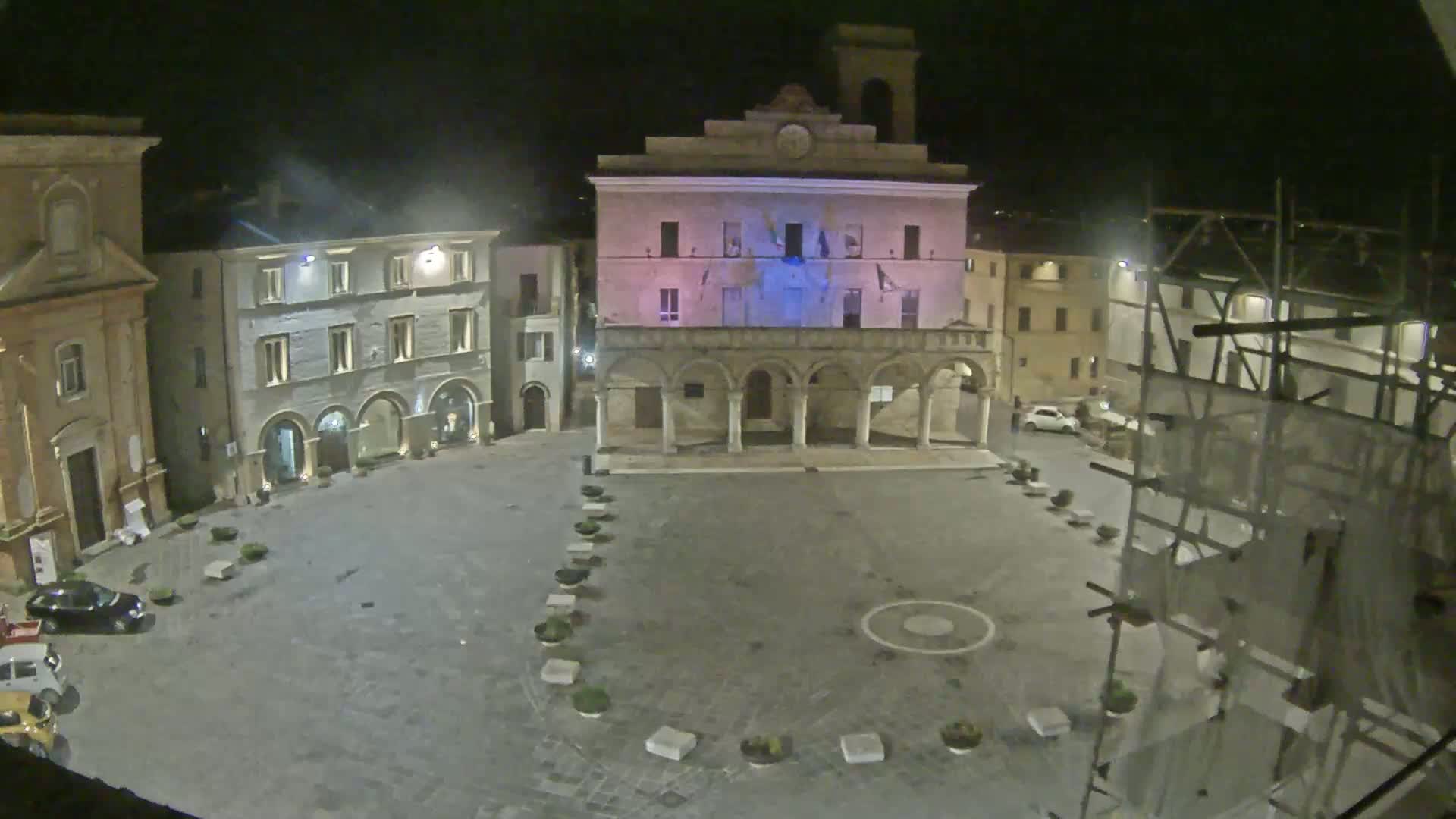 A nighttime view of a town square with several buildings, including a large building with arches, and scaffolding on one side.