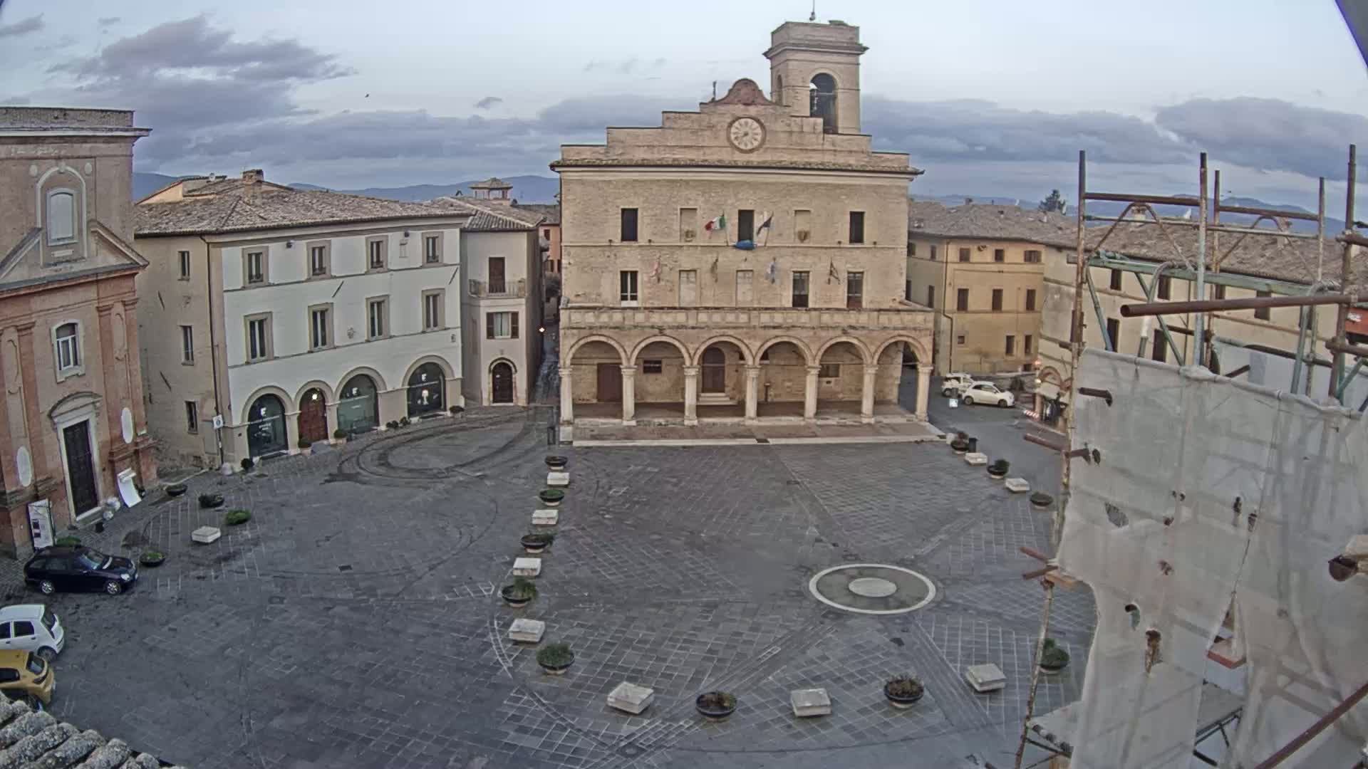 A large, mostly empty town square, under a cloudy sky, features a prominent stone building with arched entryways and a clock tower, surrounded by other buildings.