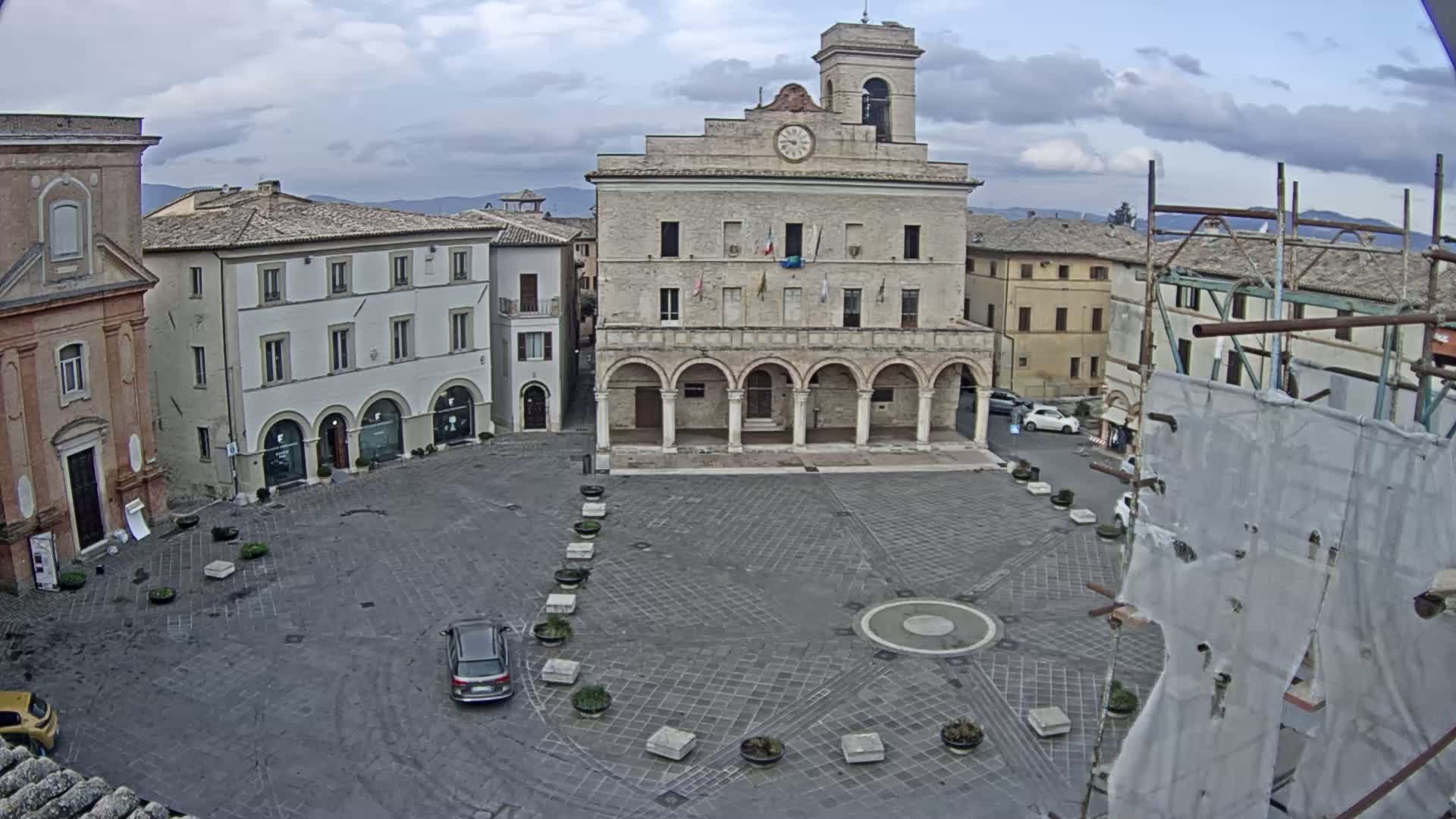 A large, stone public square, under a cloudy sky, is centered around a town hall with arched entryways, surrounded by other buildings.