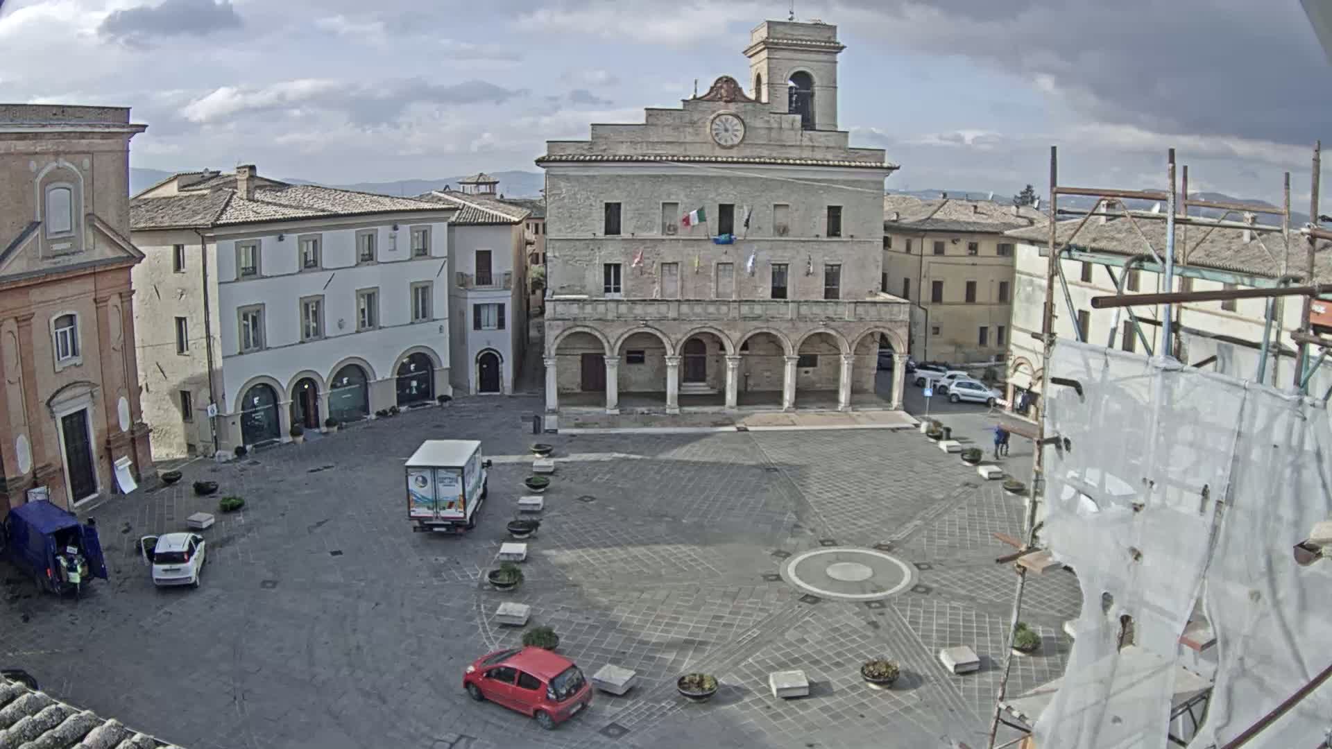 A town square with a large stone building, several cars, and a partially covered structure under cloudy skies.