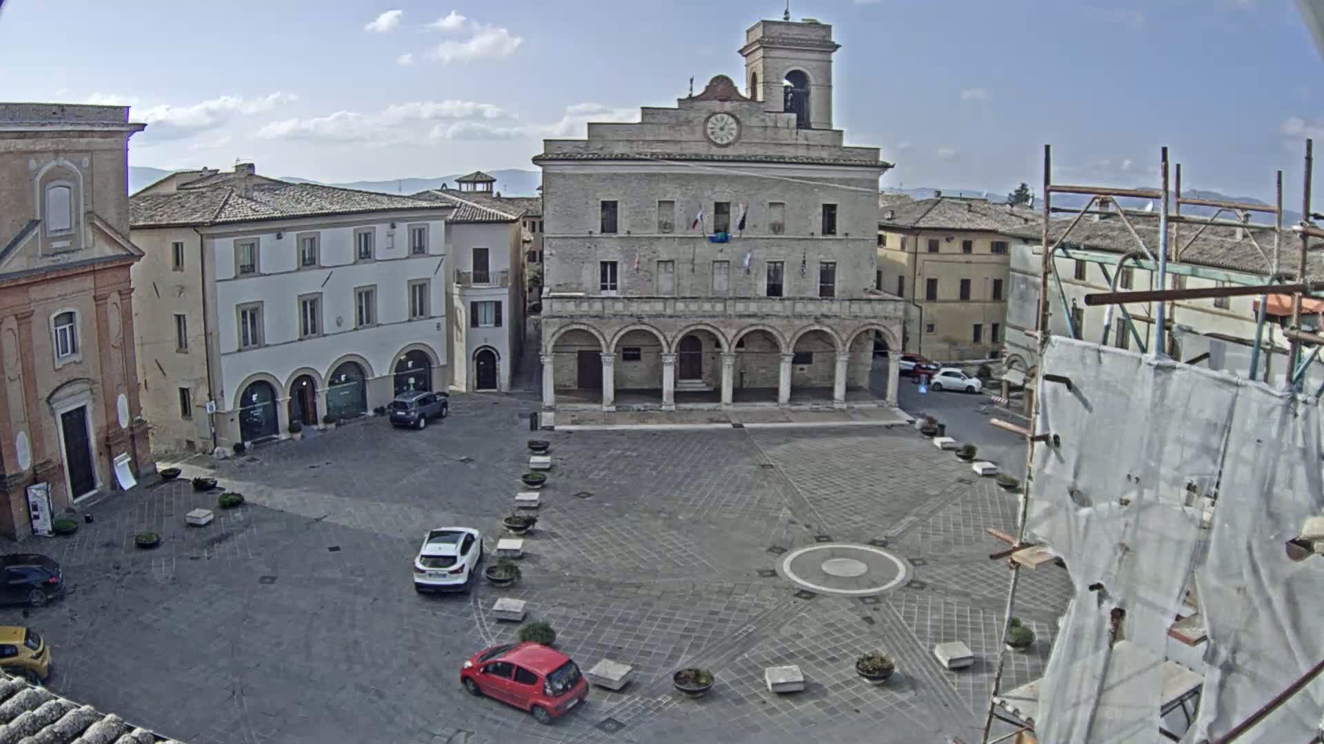 A town square with a large stone building, several smaller buildings, and a few cars parked, under a partly cloudy sky.