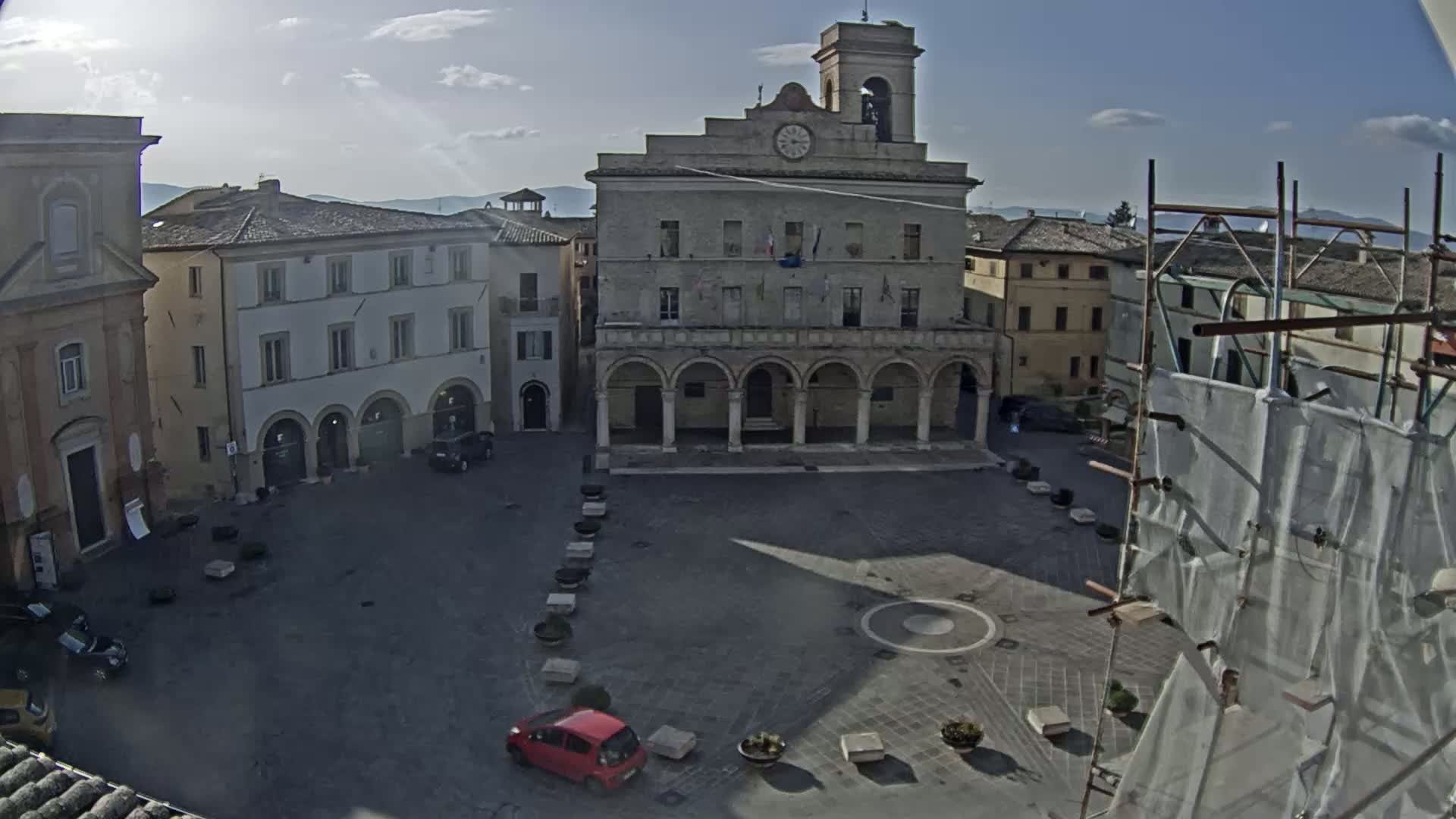 A sunny day reveals a town square with a large, arched building at its center, surrounded by other buildings, a few cars, and scaffolding.