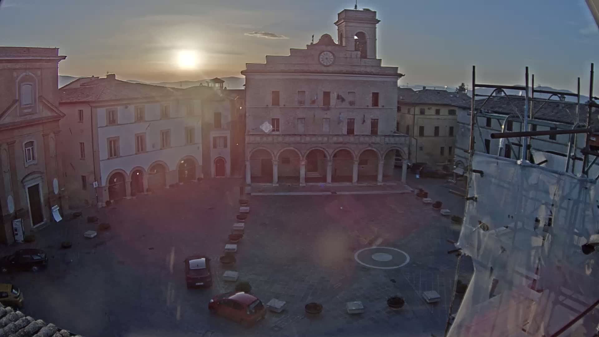 A town square at sunrise, with a large stone building featuring arched entrances at its center, surrounded by other buildings, and a few cars parked in the square.