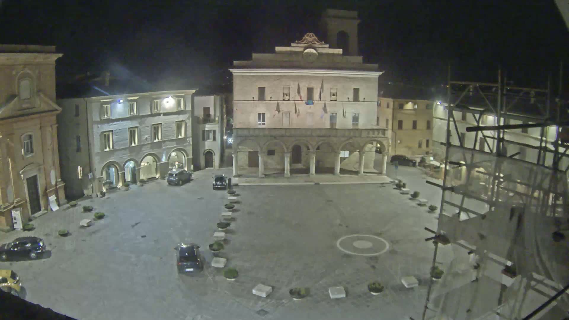 At night, a town square is illuminated, featuring a large building with arches on its ground floor, surrounded by other buildings and a few parked cars.
