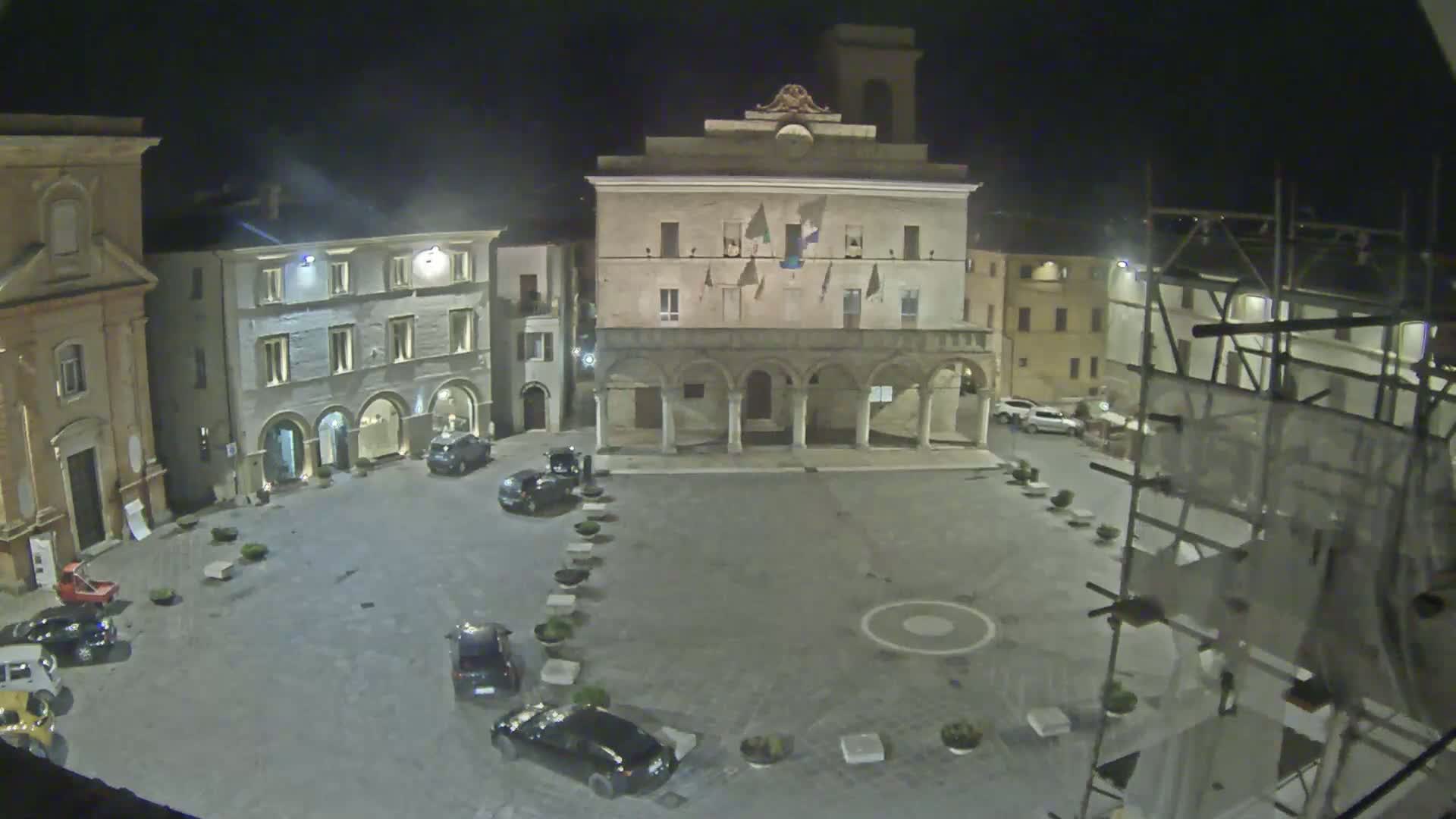 A nighttime, aerial view of a town square with several cars parked around its perimeter, centered on a large building with arched entryways.