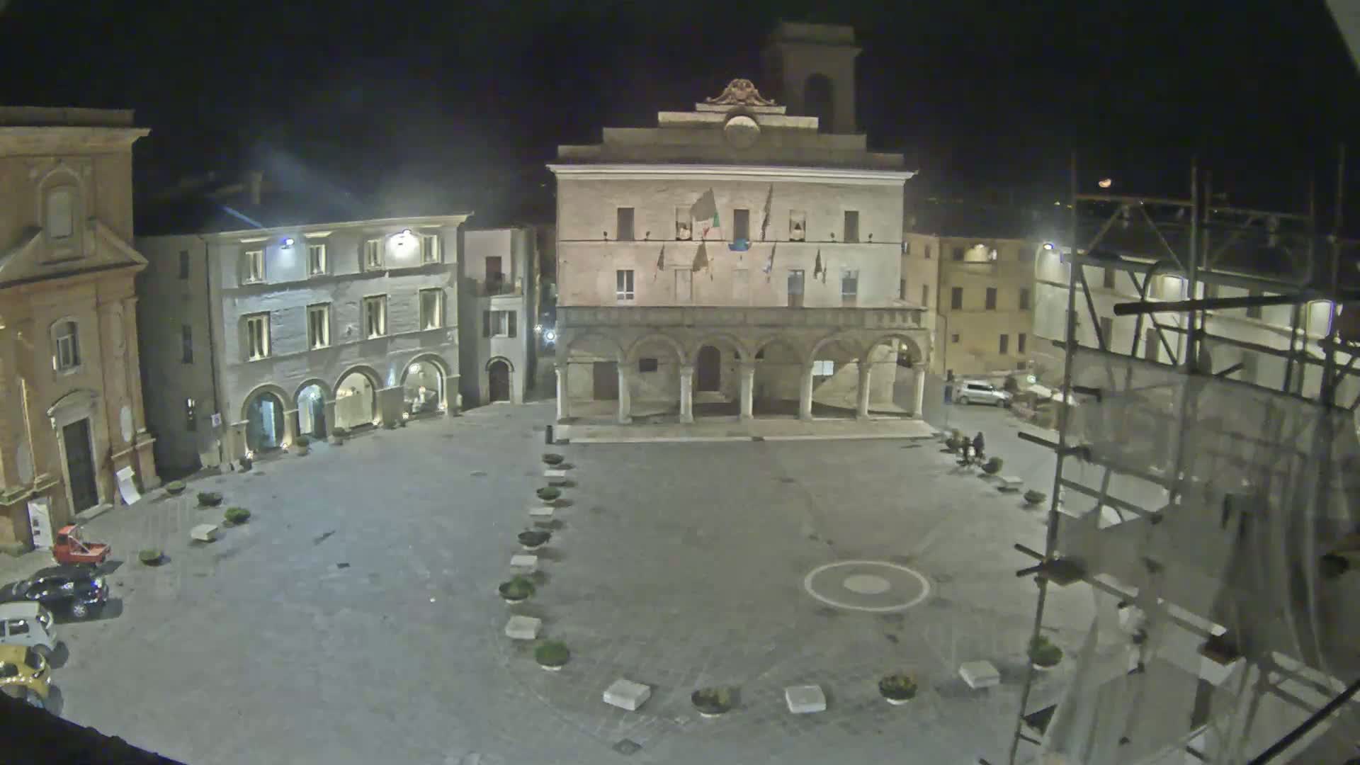 A nighttime view of a town square, featuring several buildings, including a large structure with arches, under a clear, dark sky.