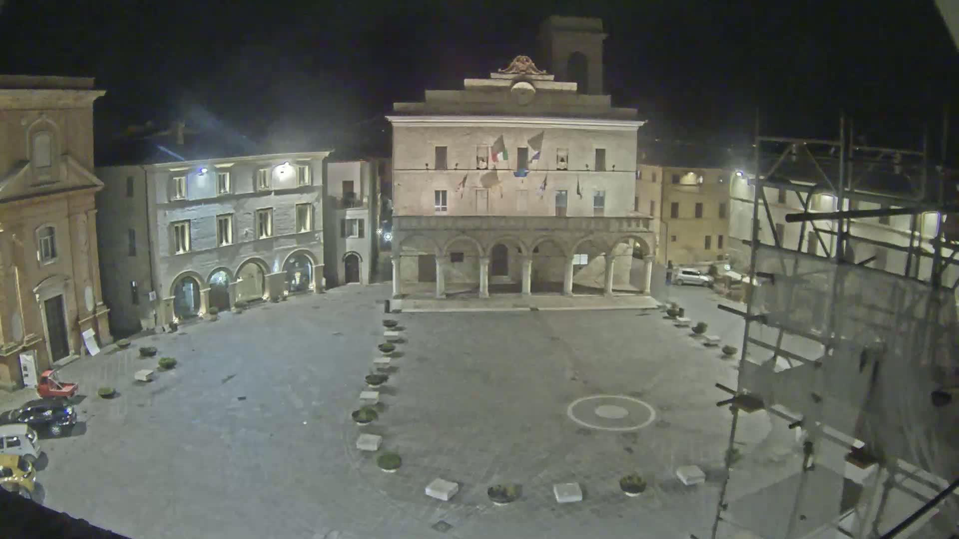 A nighttime view of a town square, featuring several buildings, a few cars, and scaffolding.