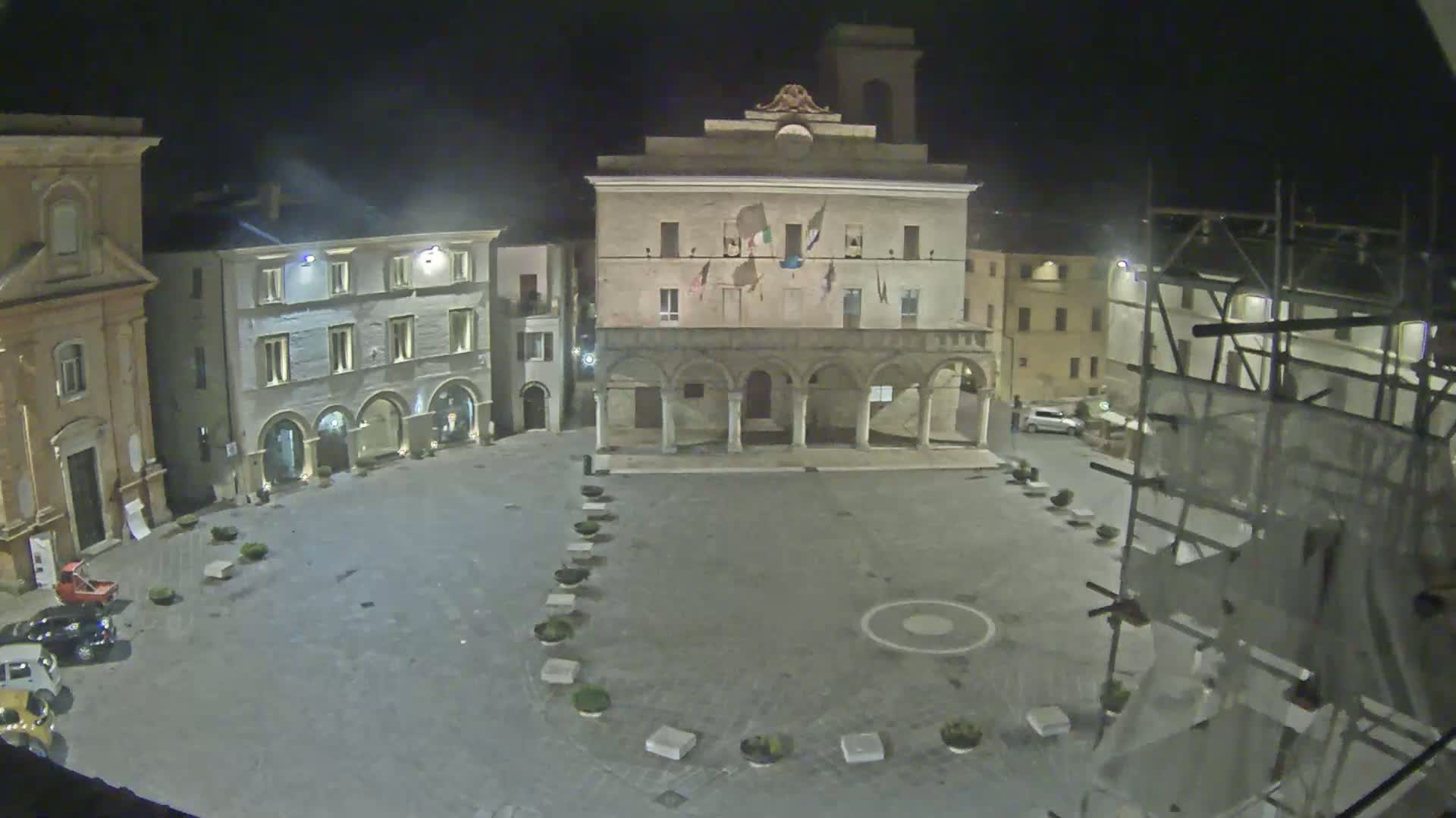 A nighttime, high-angle view of a mostly empty town square, centered on a large building with arched entryways, surrounded by other buildings.