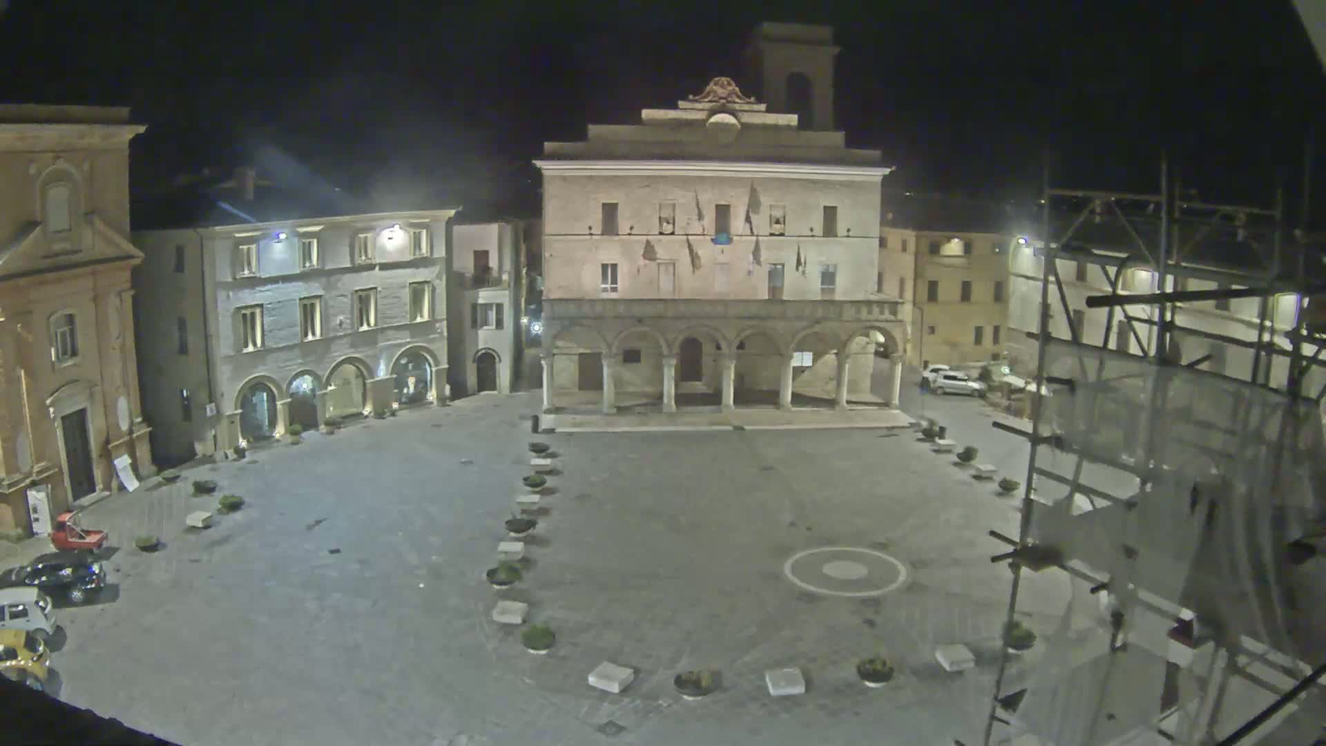 A nighttime view of a town square with several buildings, including a large building with arches, surrounded by smaller buildings and a few parked cars.