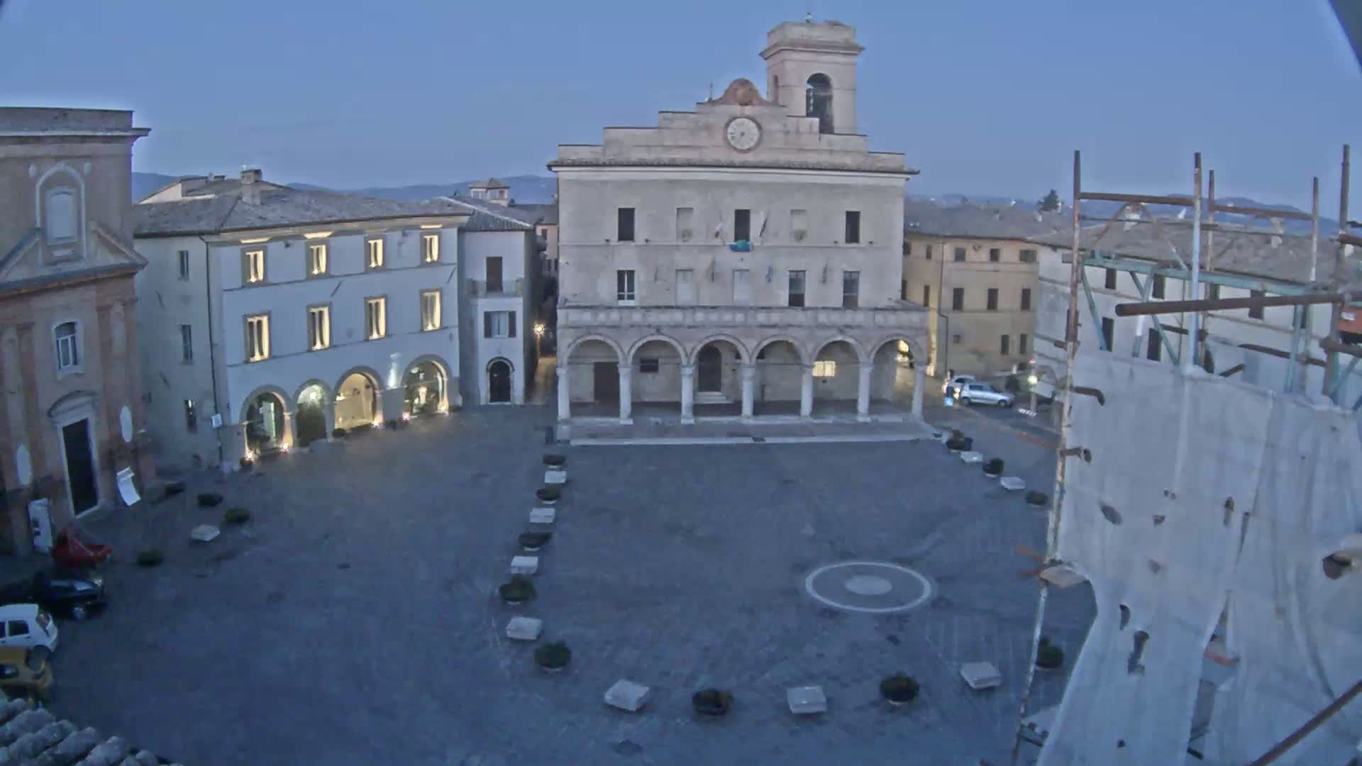 A town square at twilight is shown, featuring a large building with arched entryways, surrounded by other buildings, and a construction scaffolding in the foreground.