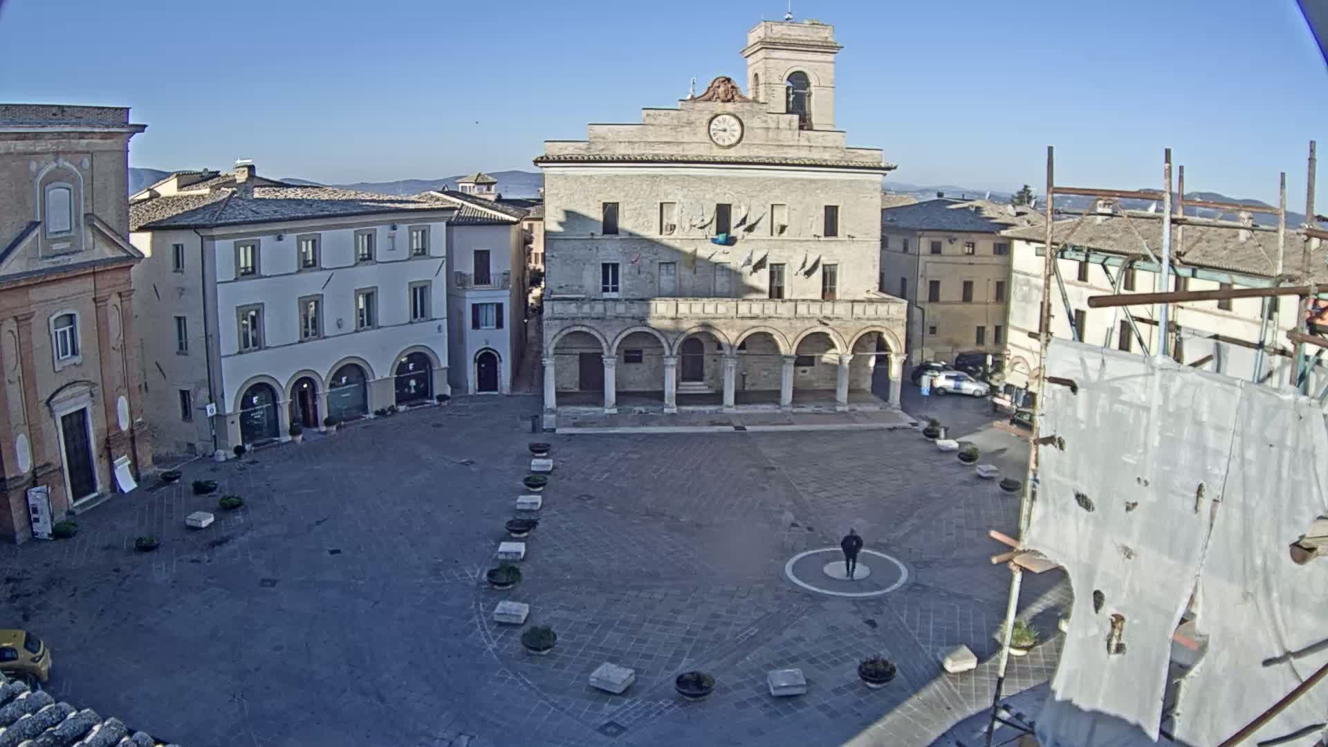 A large, mostly empty town square is surrounded by historic-looking buildings under a clear, sunny sky.