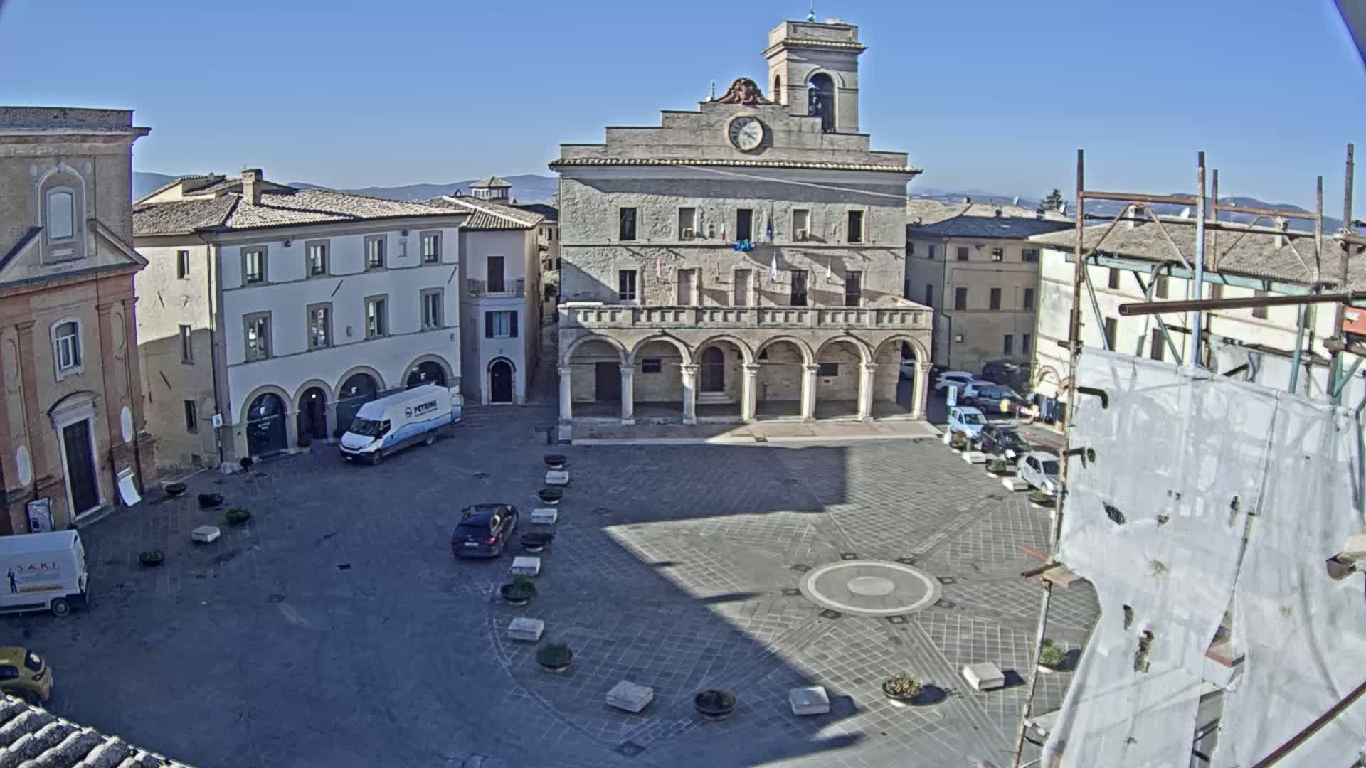 A town square with a large, light-colored building in the center, surrounded by other buildings, under a clear, sunny sky.