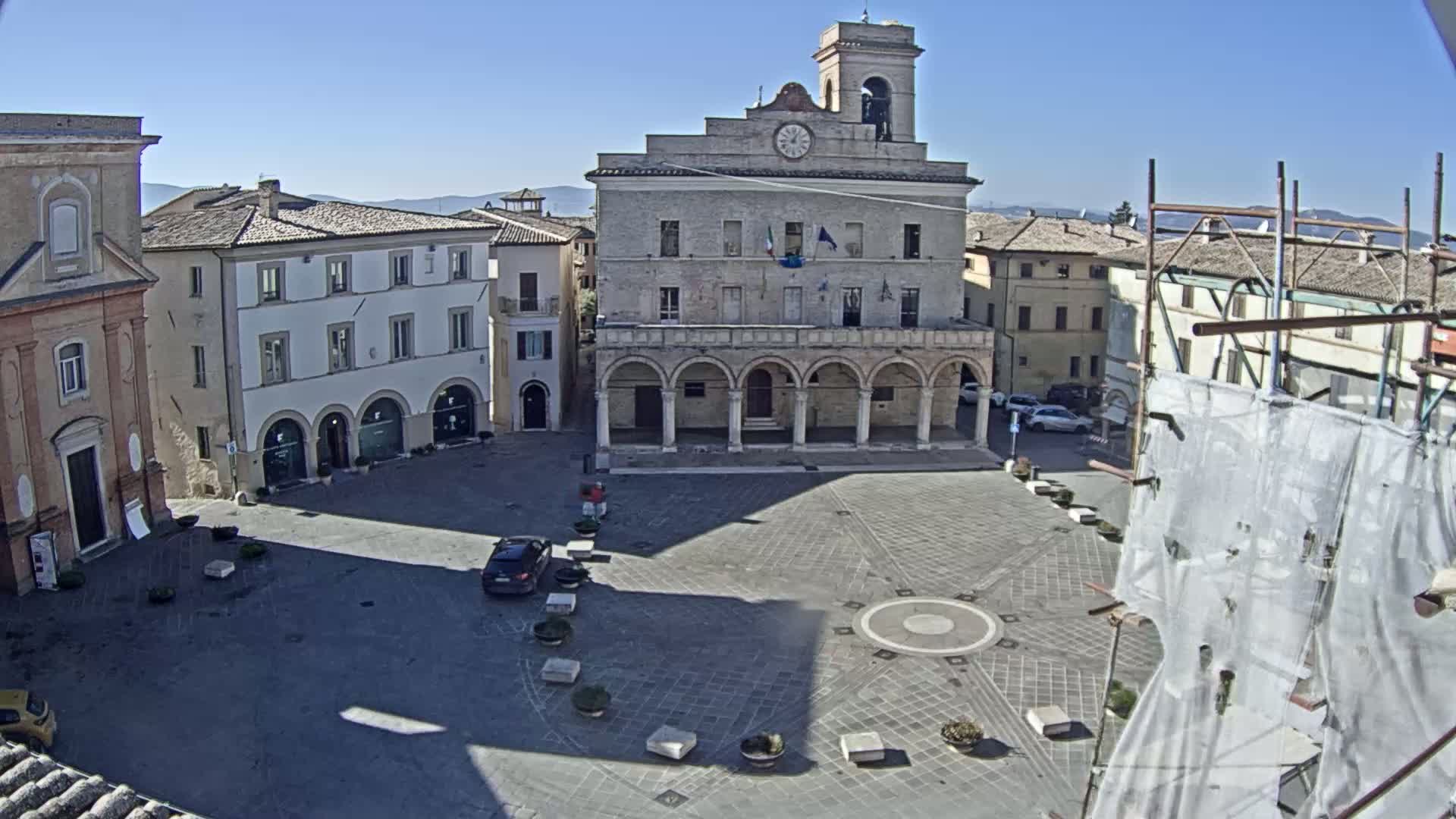 A town square, under sunny skies, features a large stone building with arched entryways, surrounded by other buildings, a few cars, and several potted plants.