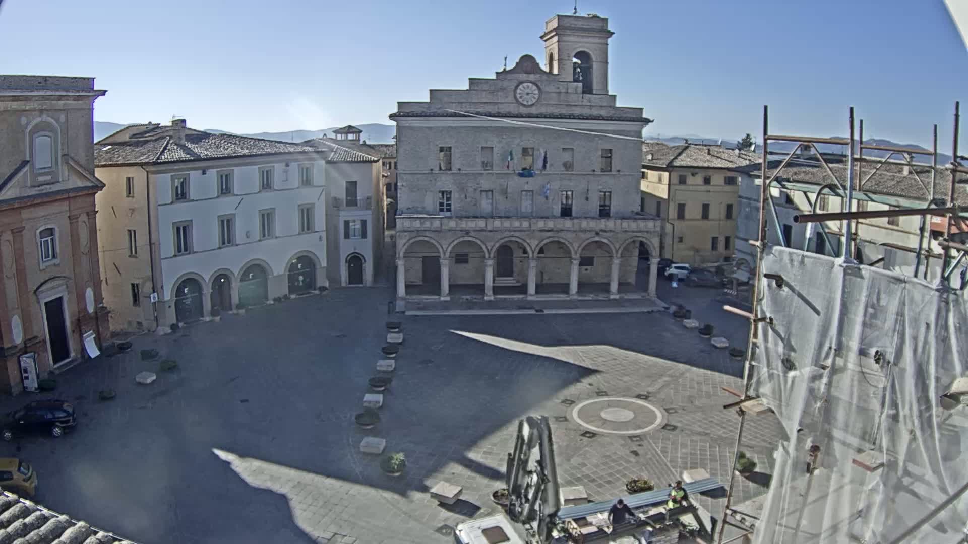 A sunny day reveals a town square, centered around a large, arched building with a clock tower, surrounded by other buildings.