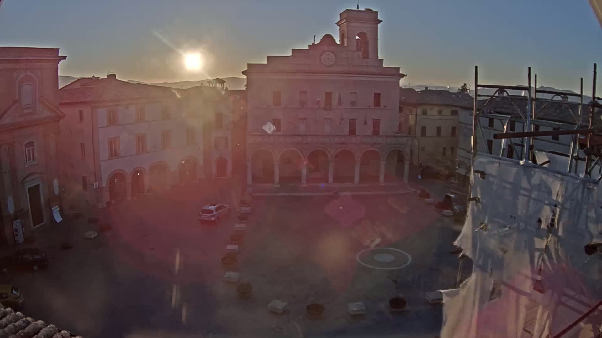 A sunlit town square with several buildings, including a large stone building with arched entryways, is seen at sunrise.
