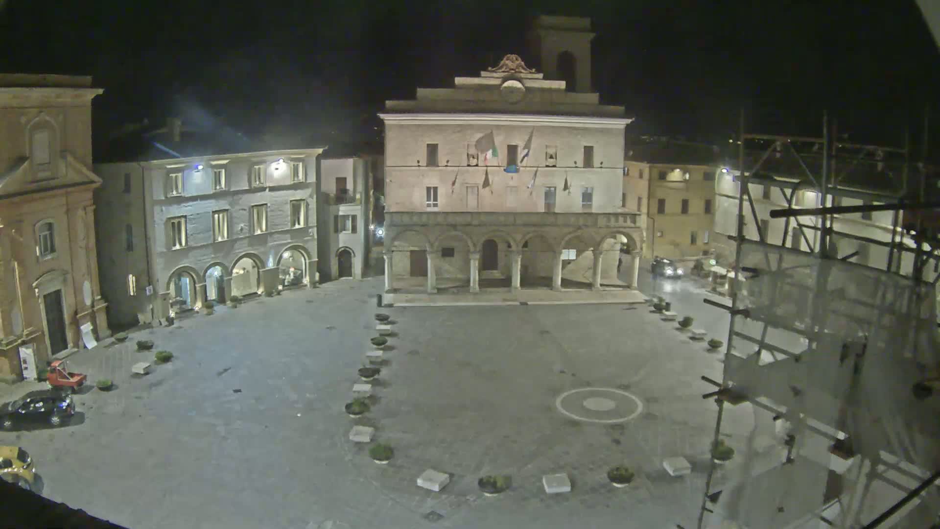 A nighttime, dimly lit town square, dominated by a large, arcaded building, with several smaller buildings surrounding it and scaffolding on one side.