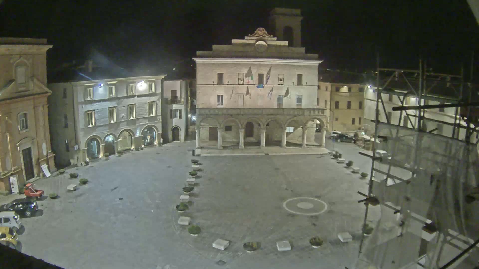 A nighttime view of a town square with several buildings, including a large building with arched entryways, under a dark sky.