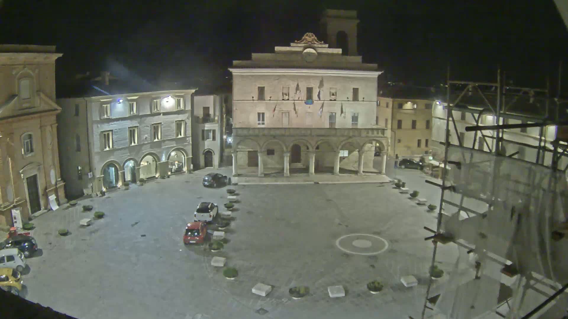 A nighttime, aerial view of a town square with several cars parked, surrounded by historic buildings, one of which has arched entrances and flags hanging from it.