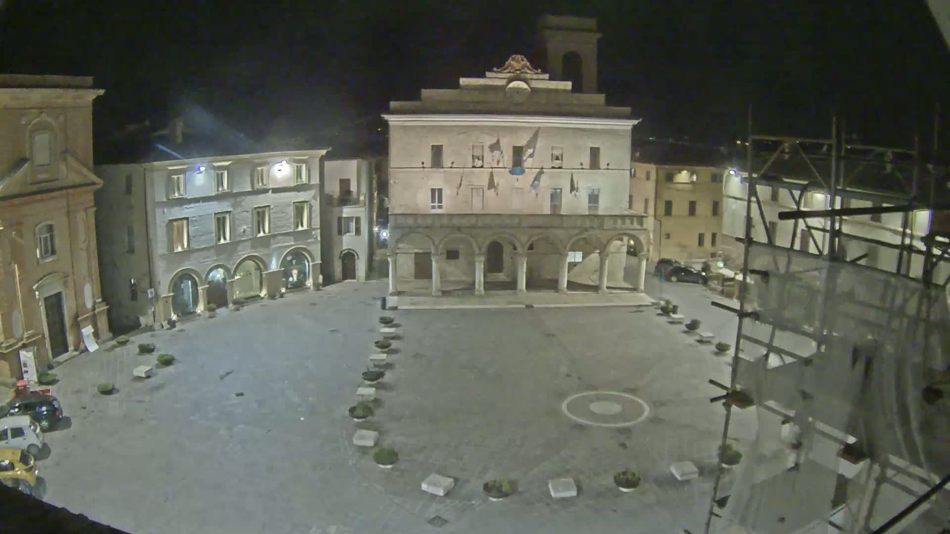 An empty town square at night, illuminated by artificial light, is surrounded by buildings, including a large, arched structure, and scaffolding is visible on one side.