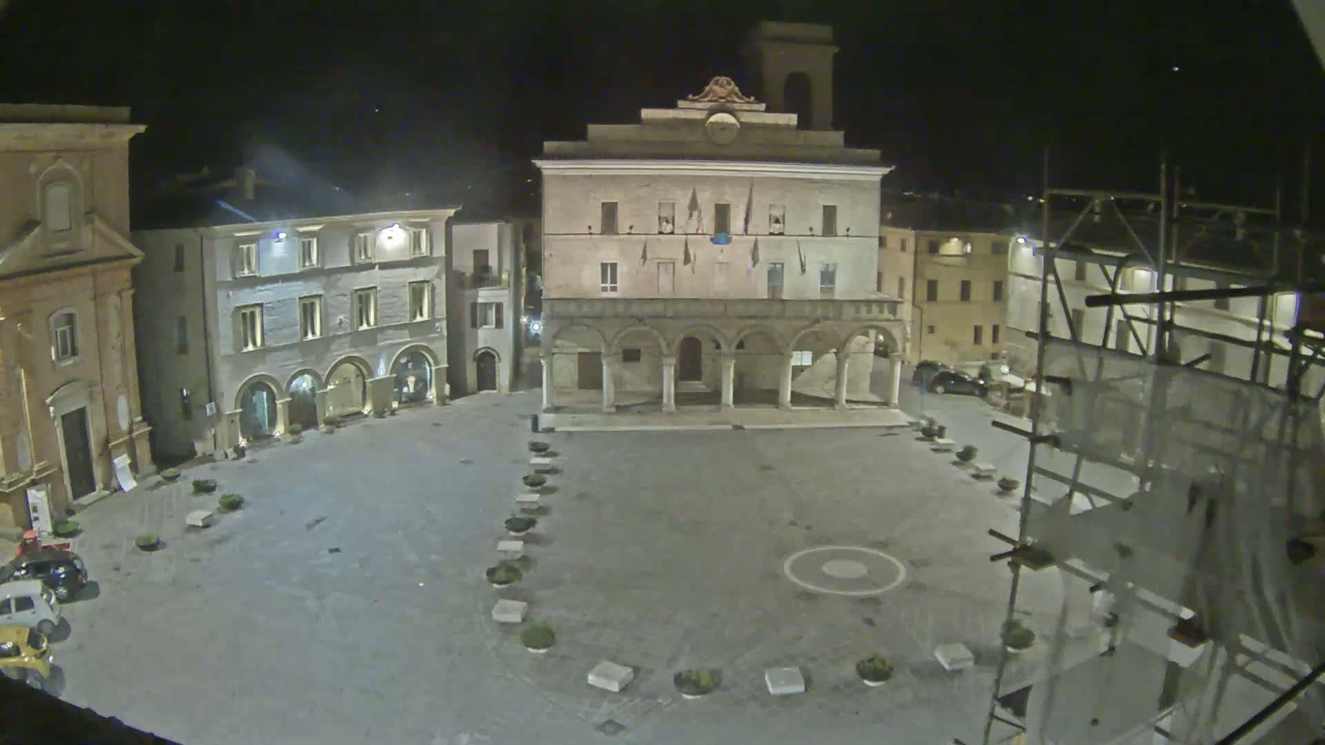 A nighttime view of a town square, featuring a large building with arches on the ground floor, surrounded by other buildings, under a clear, dark sky.