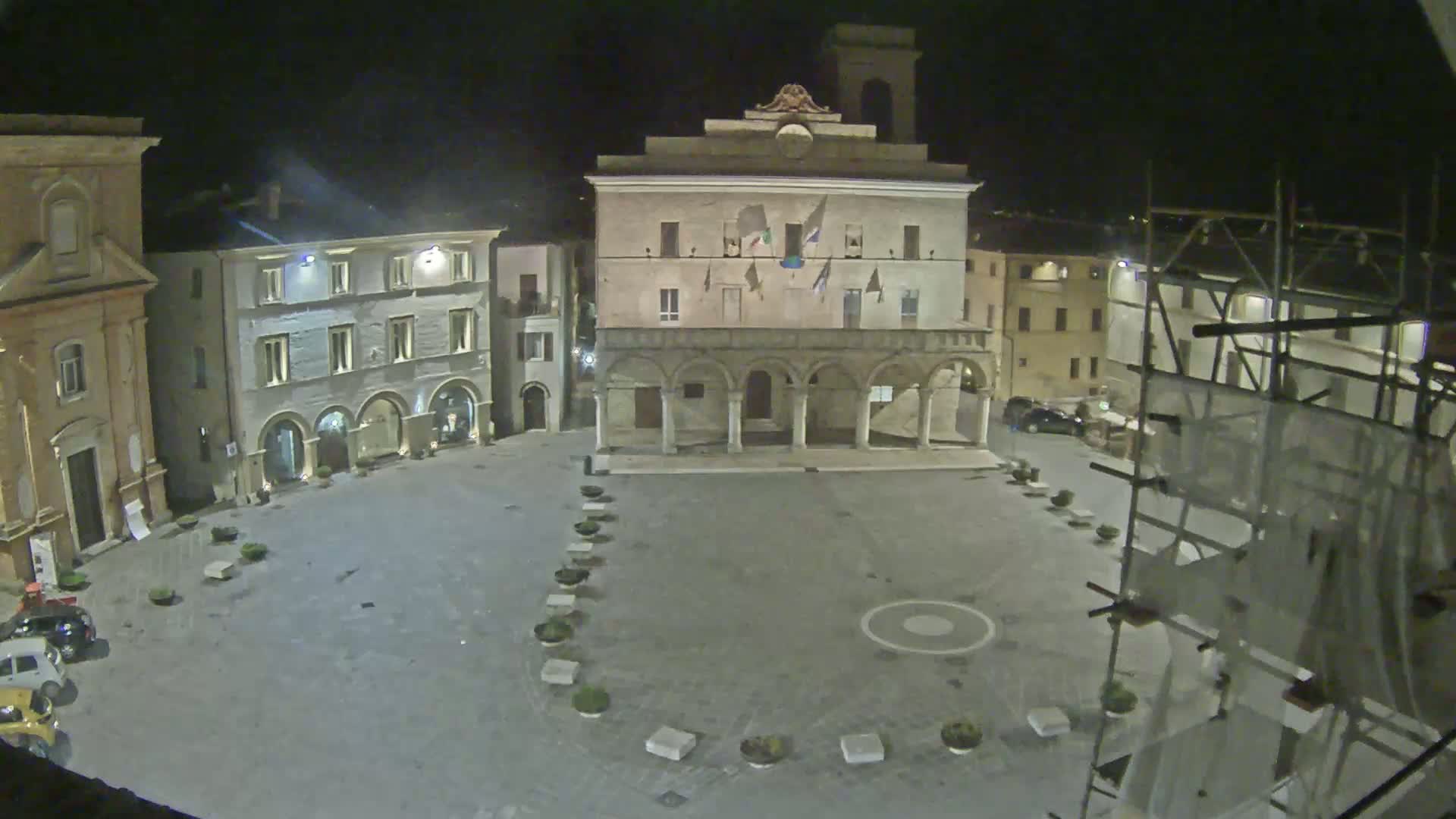 A nighttime, high-angle view of a town square, featuring several historic buildings, a few parked cars, and scaffolding on one side.