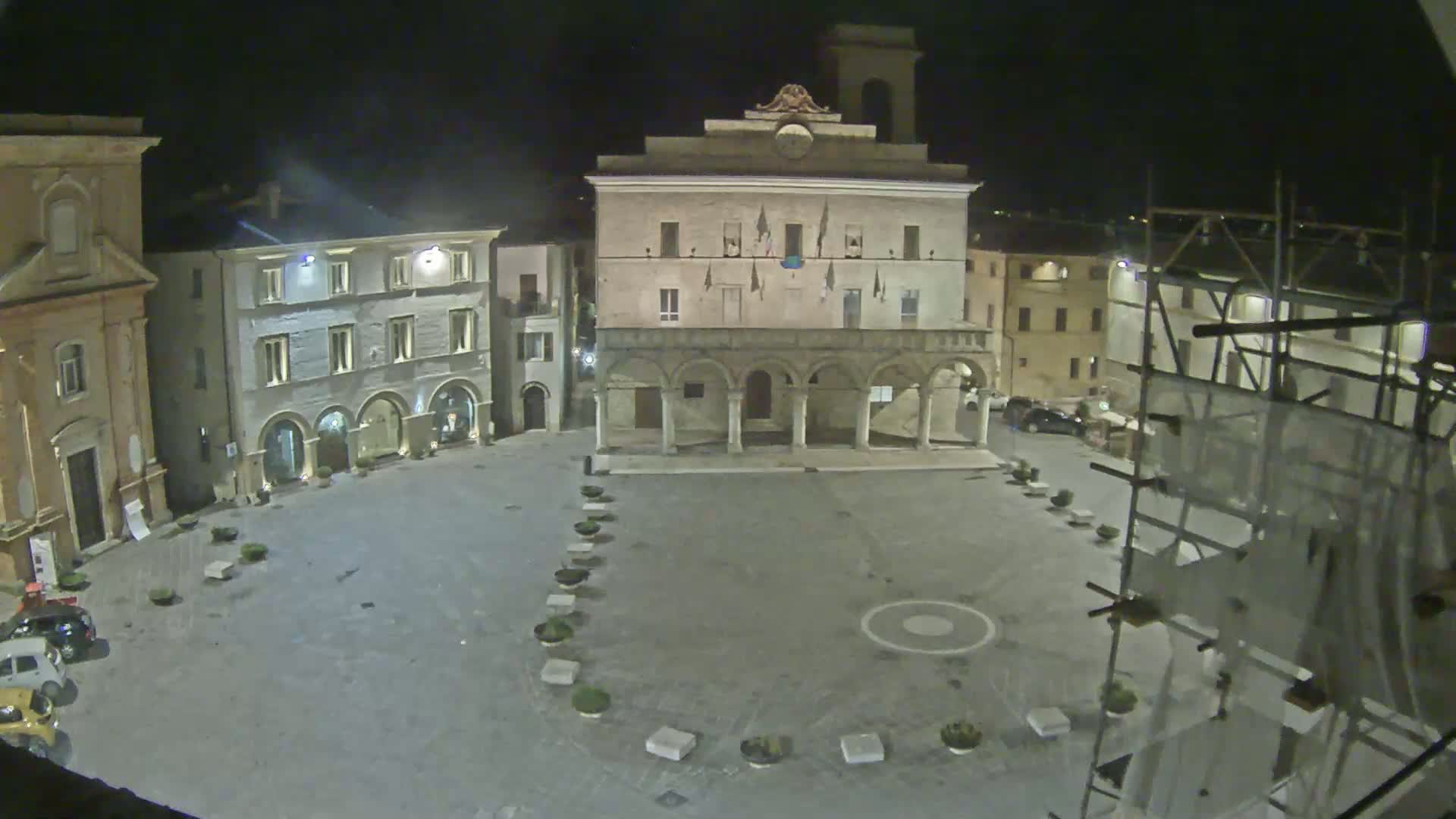An empty town square at night, illuminated, is surrounded by historic-looking buildings, one of which is under construction.