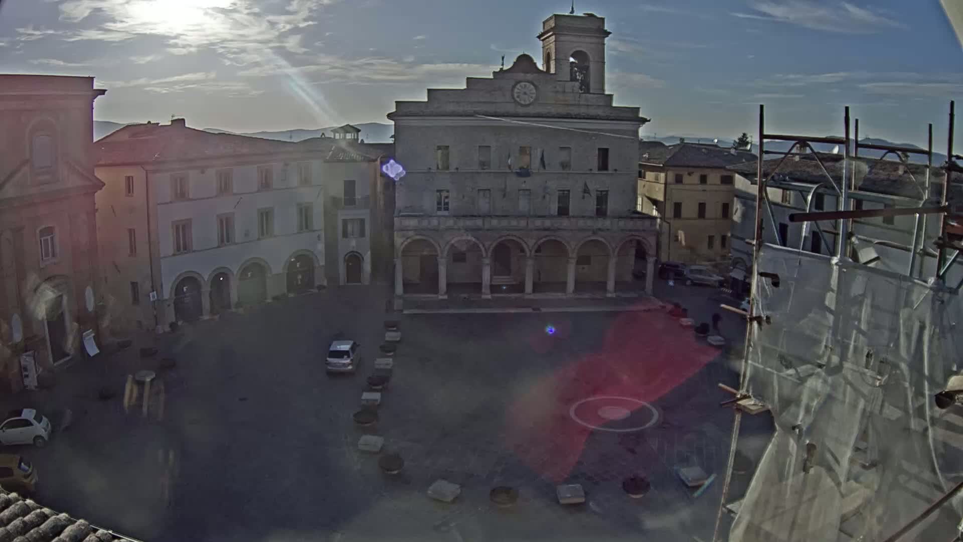 A sunny day reveals a town square, centered around a large, classical-style building with arched entryways, surrounded by other buildings, with a few cars parked in the square.