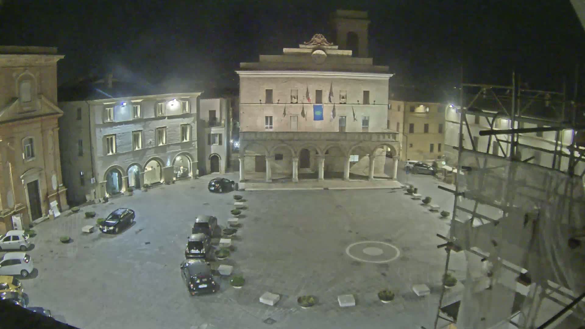 A nighttime view of a town square with several cars parked, surrounded by buildings, one of which has a colonnade.