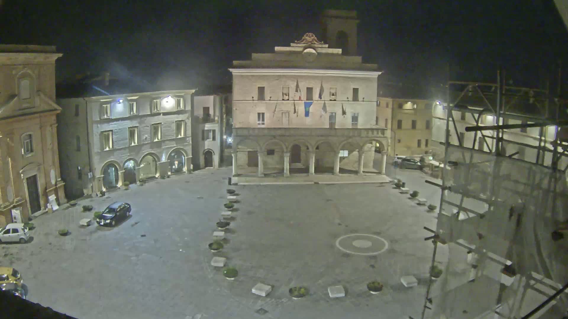 An empty town square at night, illuminated by artificial light, is surrounded by historical buildings, one of which is under construction.