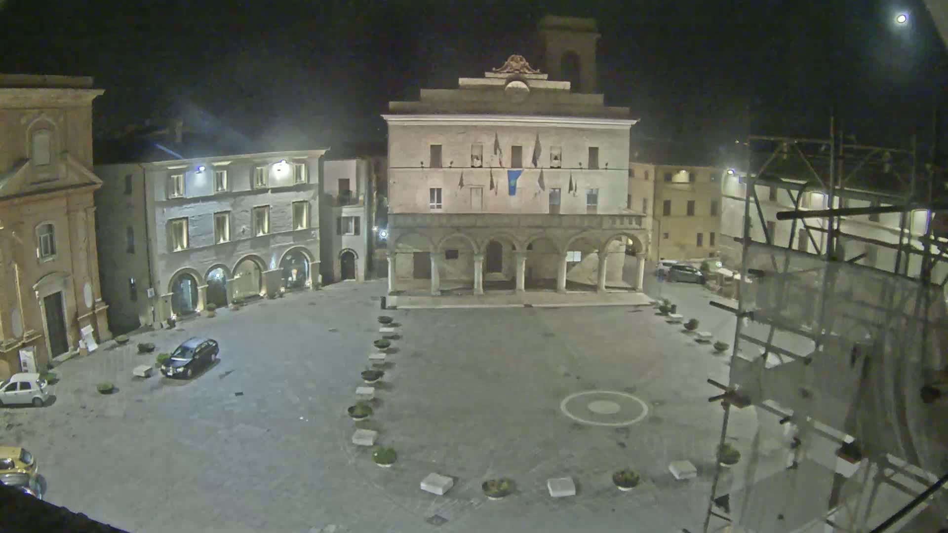 A nighttime view of a town square, featuring several buildings, a few parked cars, and scaffolding, under a mostly clear sky.