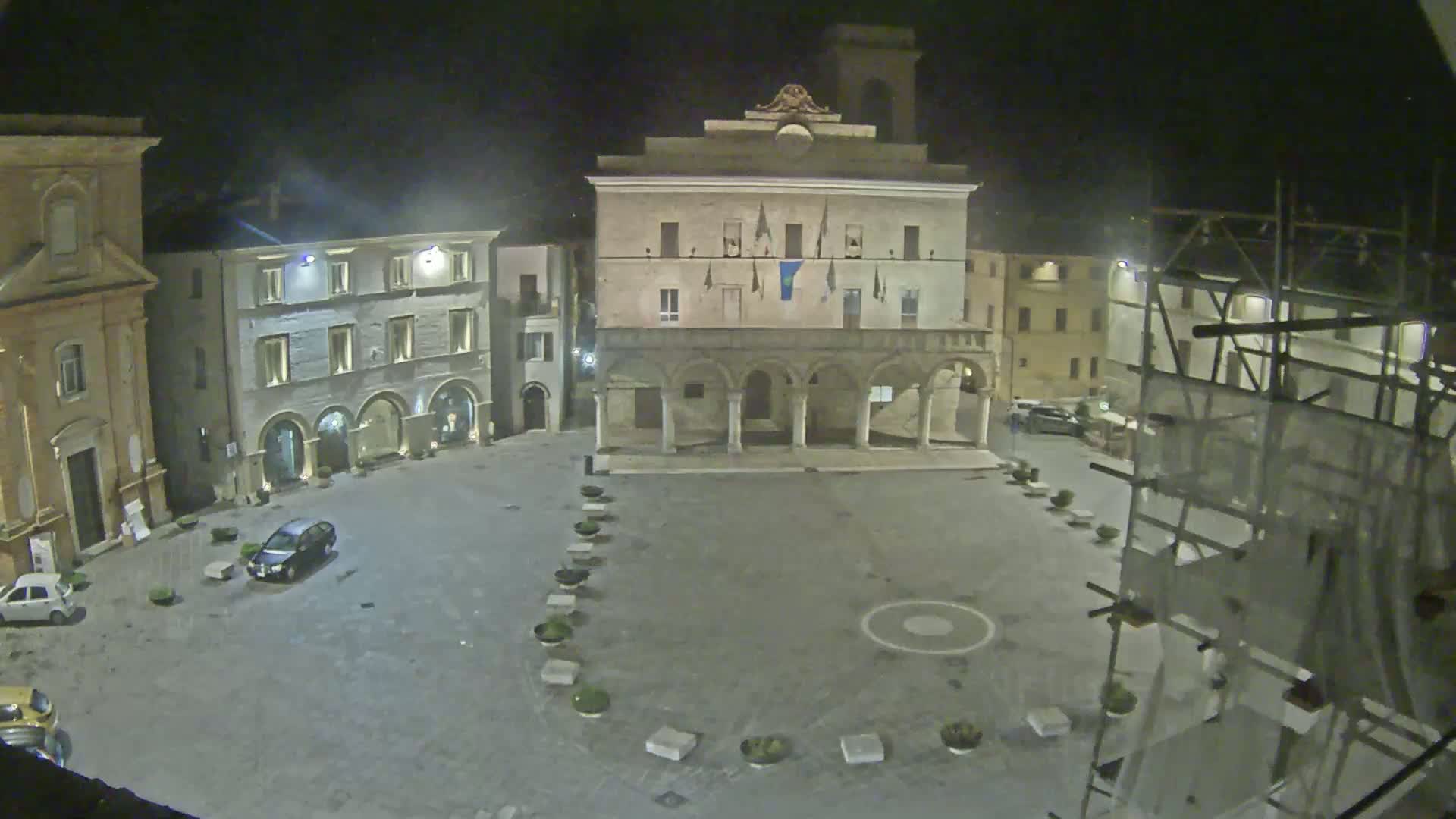 A mostly empty town square at night is surrounded by historic buildings, one of which is under construction.