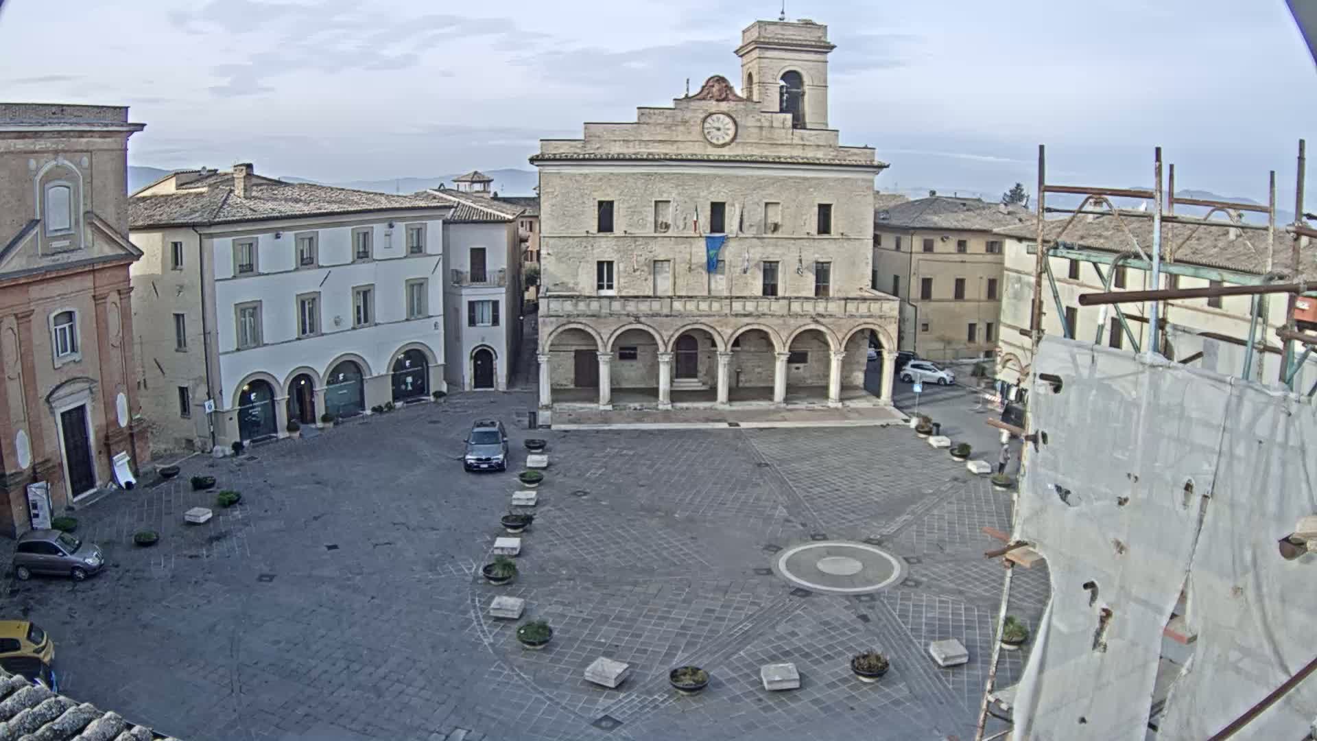 A large town square with a clock tower and several buildings surrounding it, under a mostly cloudy sky.
