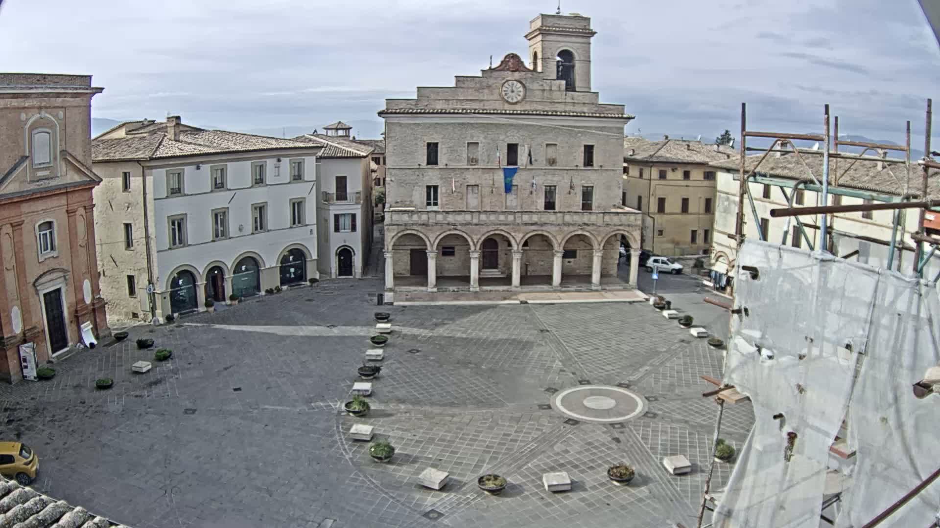 A large town square with a clock tower and several buildings surrounding it under a partly cloudy sky.