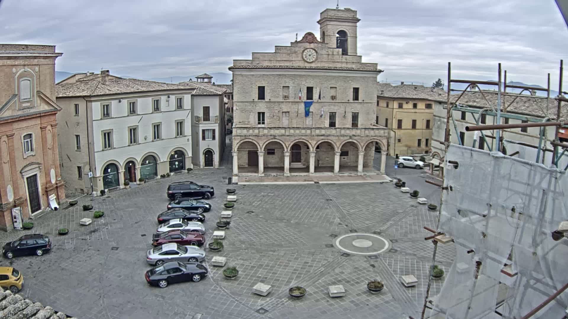 A town square with several cars parked in a row, surrounded by historical buildings under an overcast sky.