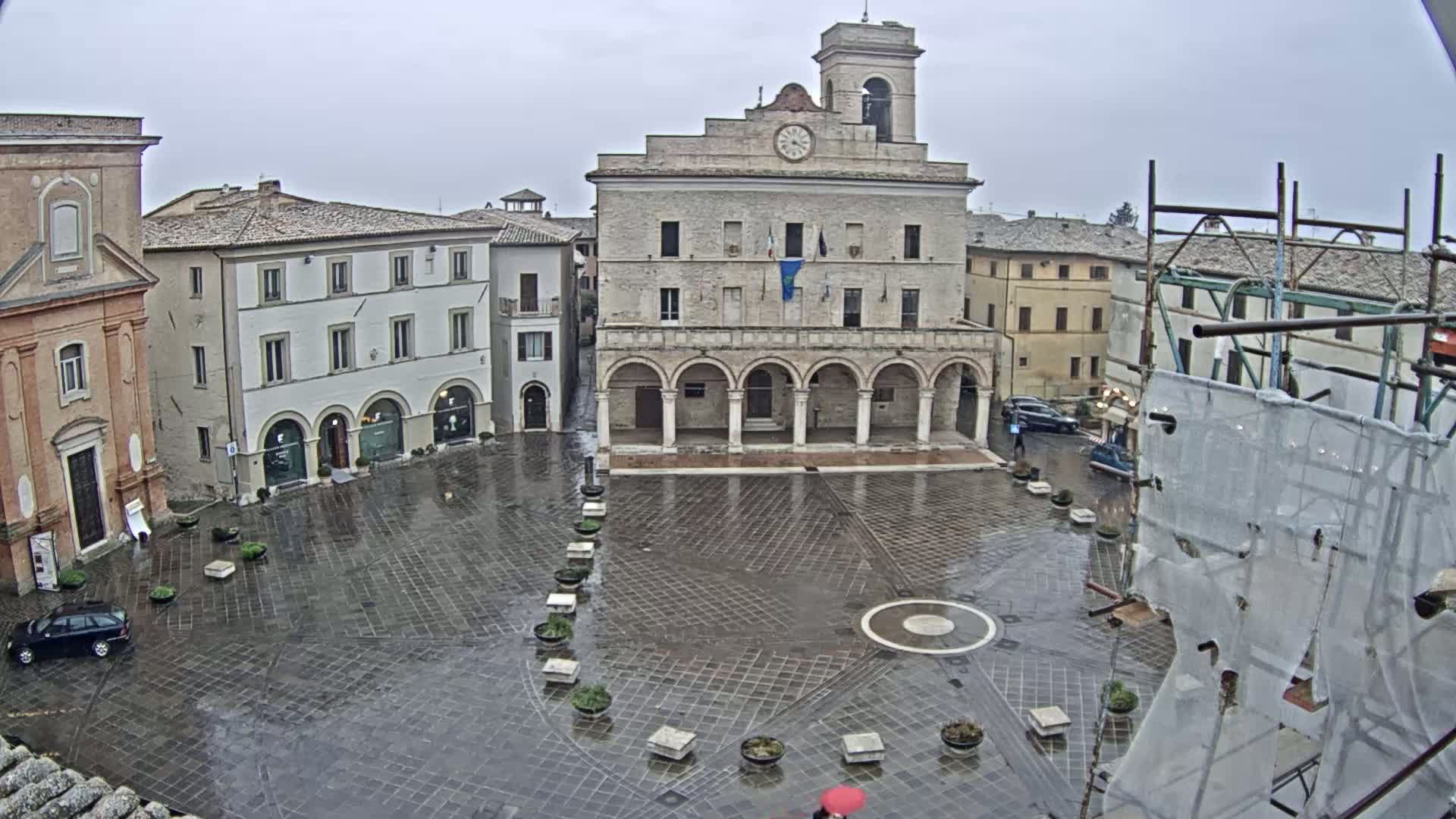 A wet, paved town square is centered on a large, arched building, surrounded by other buildings under an overcast sky.