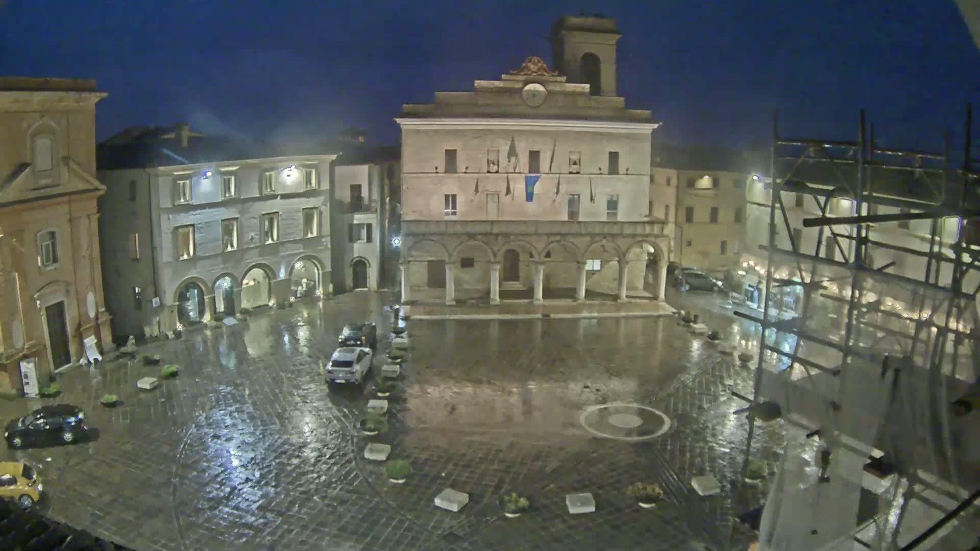 A rain-slicked town square at night, centered on a large, arcaded building, surrounded by other buildings under a dark sky.